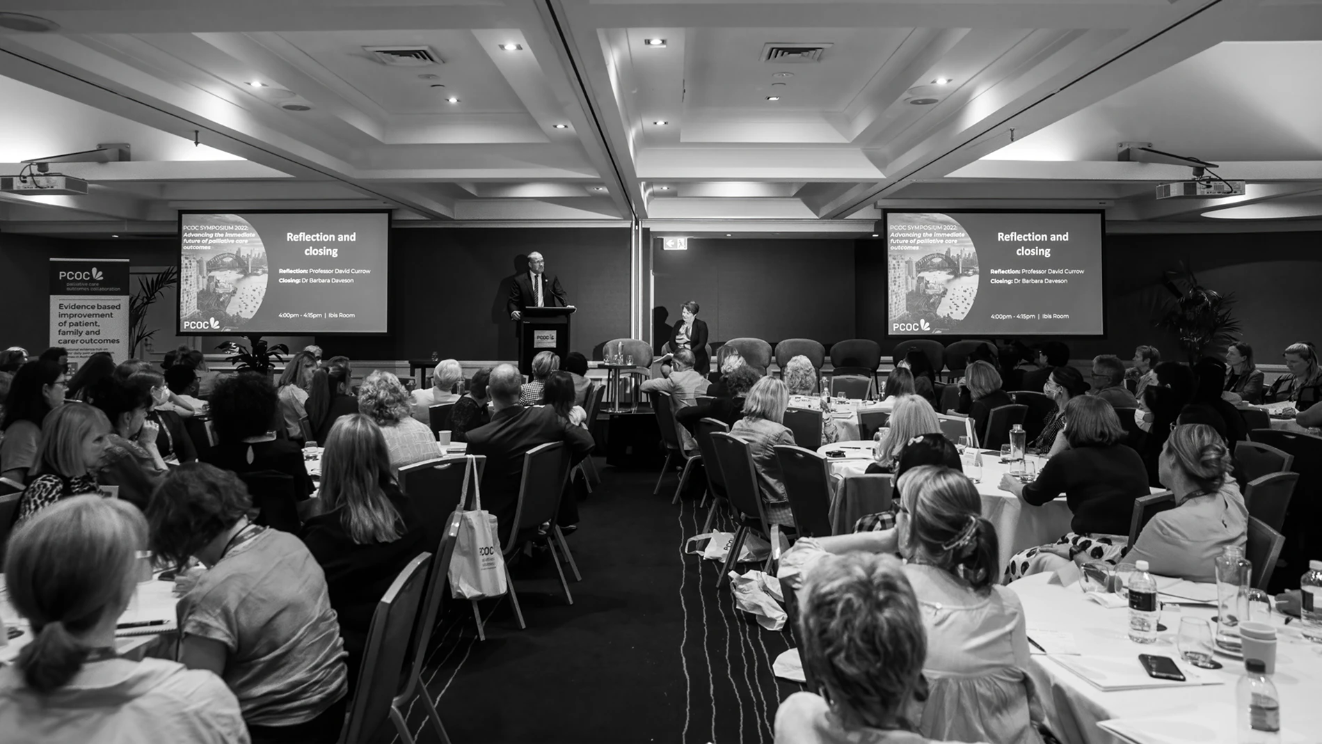 The Palliative Care Outcomes Collaboration Symposium 1 Conference Room With Attendees At Round Tables, Speaker At Podium, Projected Slides Visible.