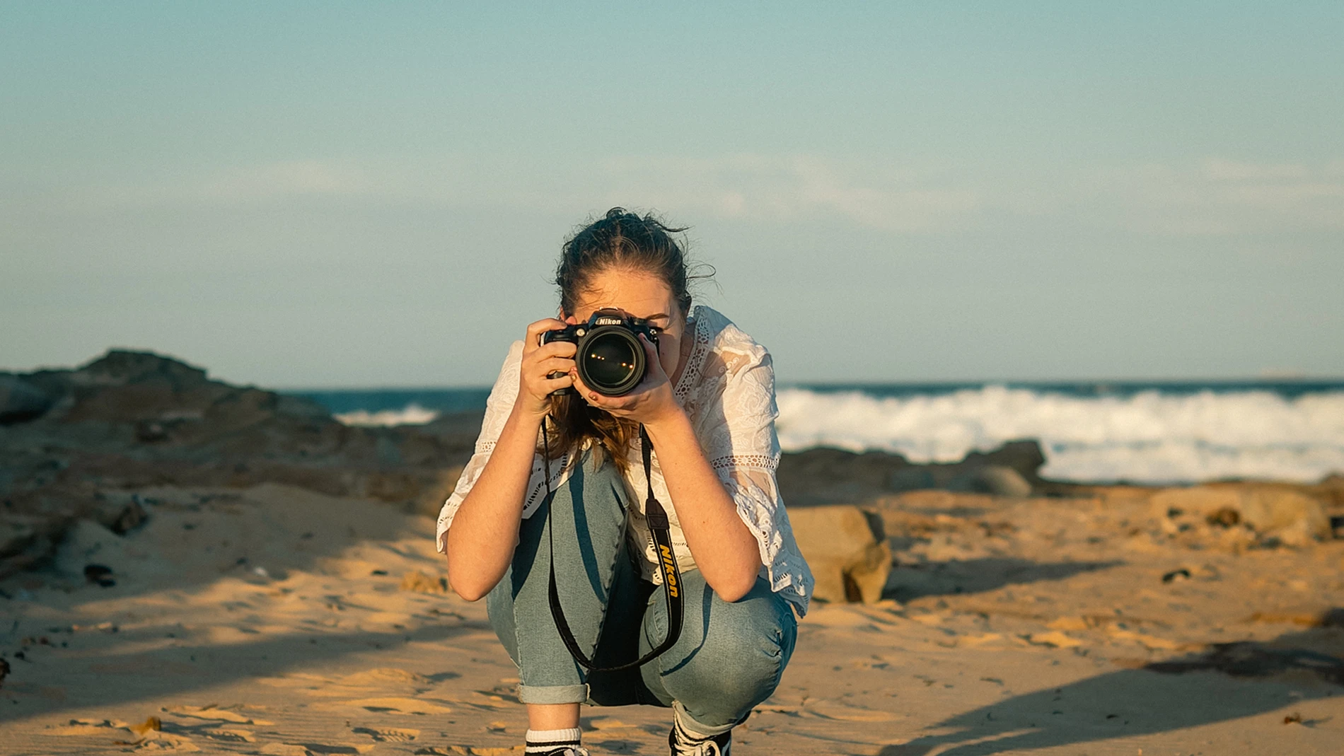 The Best Photography Locations In Wollongong 1 Person Crouching On Sandy Beach With Camera, Waves In Background, Wearing Casual Clothes.