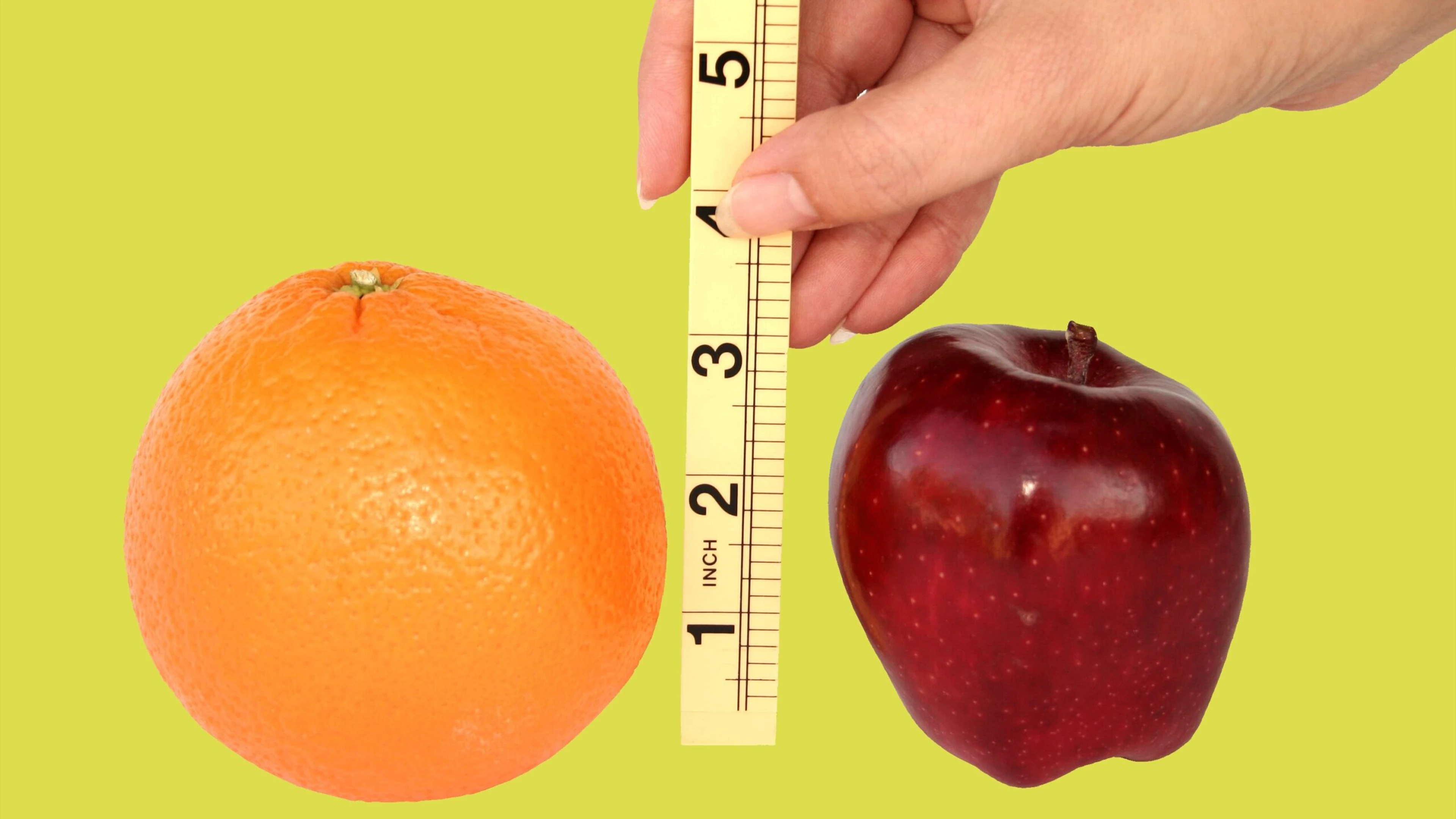 Orange and red apple side by side with a vertical ruler between them on a light green background.