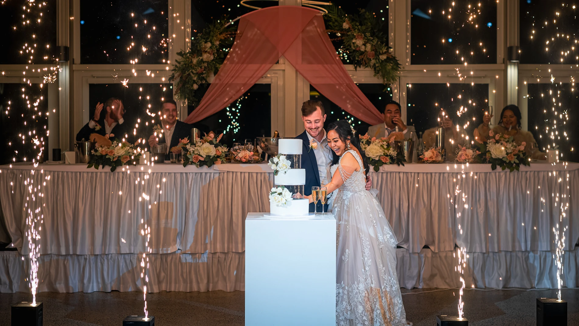 Couple cutting wedding cake amidst fireworks, with bridal party and elegant decor in the background.