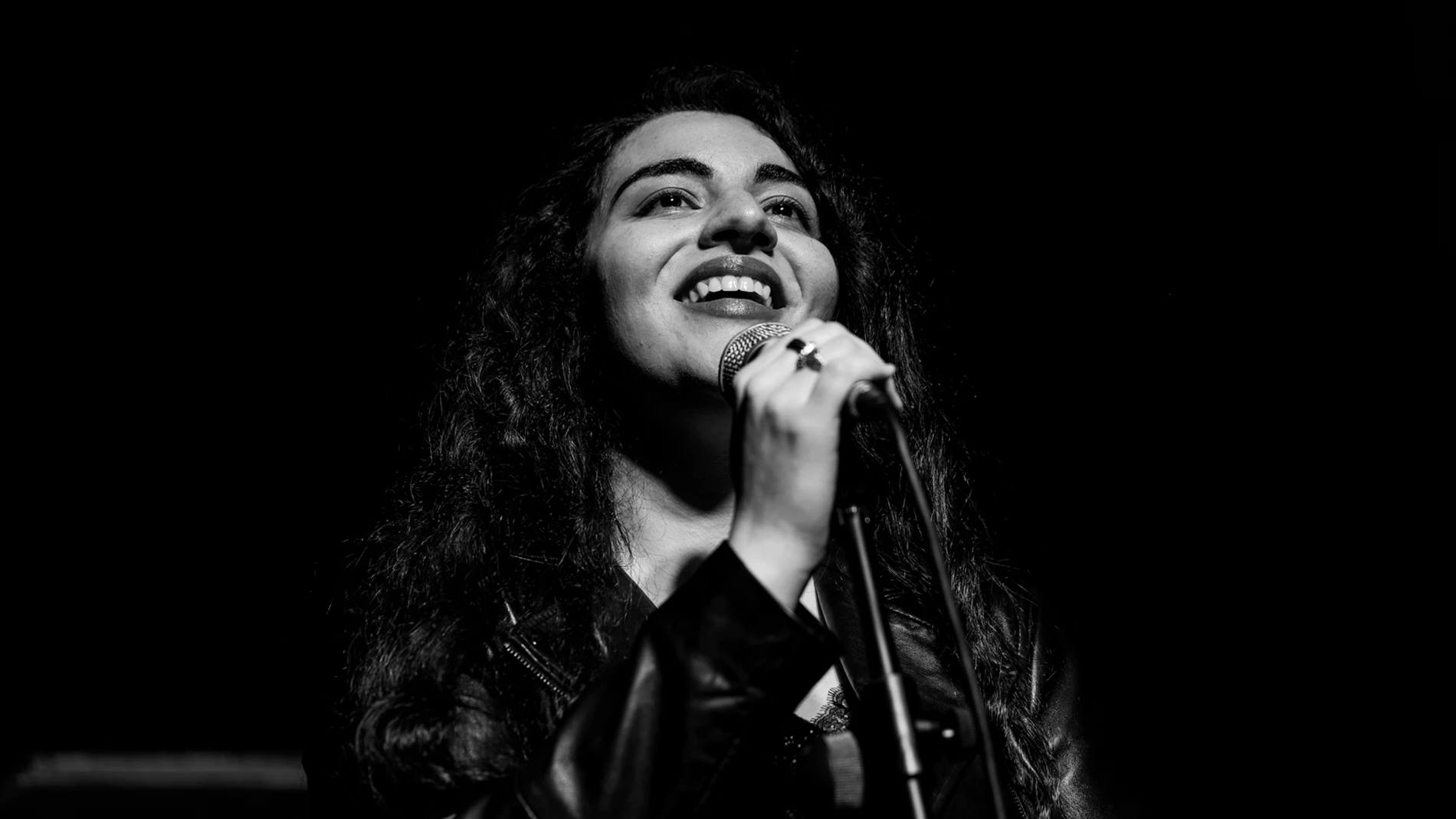 Black And White Photo Of Person With Curly Hair Singing Into A Microphone, Wearing A Dark Jacket.