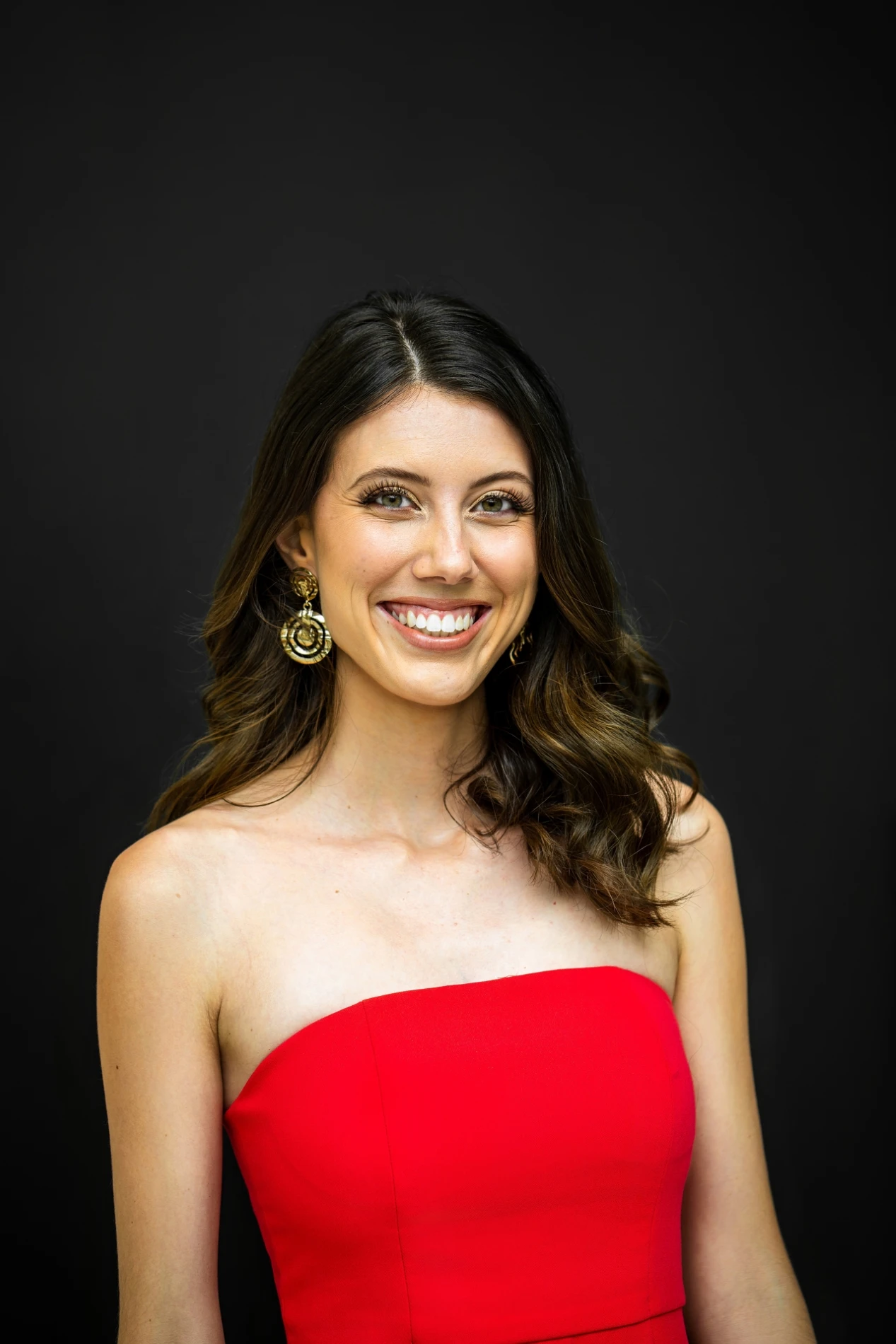 Smiling Person With Long Hair, Red Strapless Dress, Circular Earrings, Dark Background.