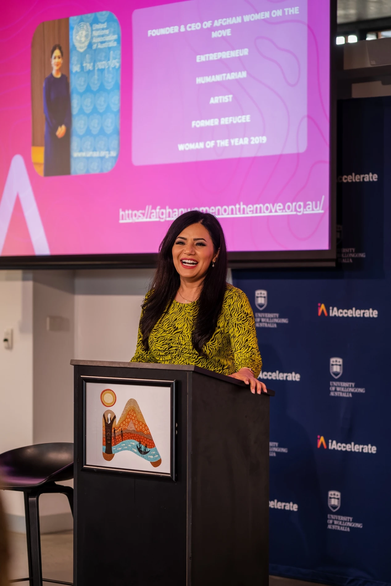 Woman Presenting At Podium With Slide On Achievements, Iaccelerate Banner In Background, Uow Australia.