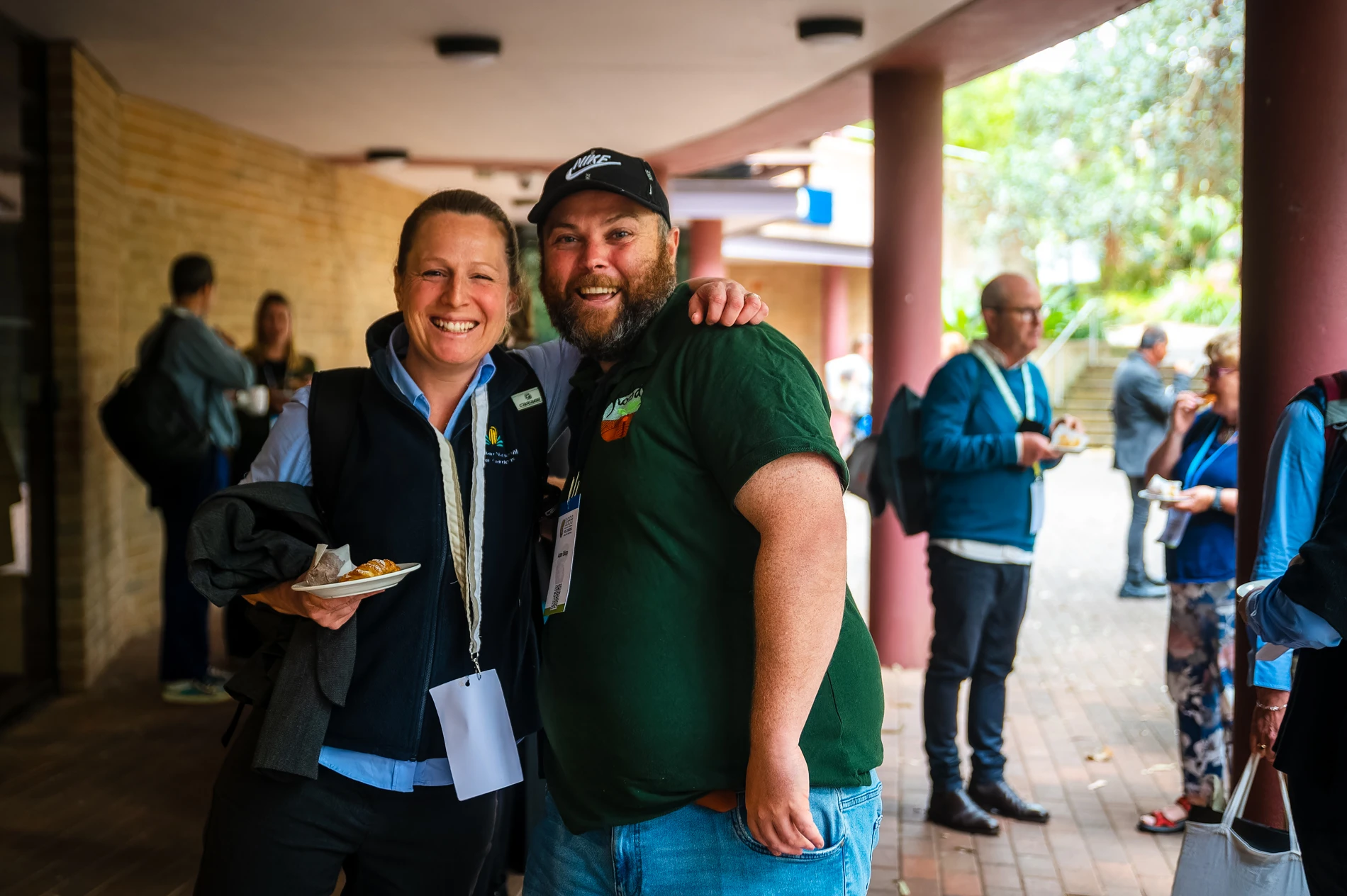 Smiling Duo Pose At Outdoor Event, Others Mingle With Food And Drinks In Background.