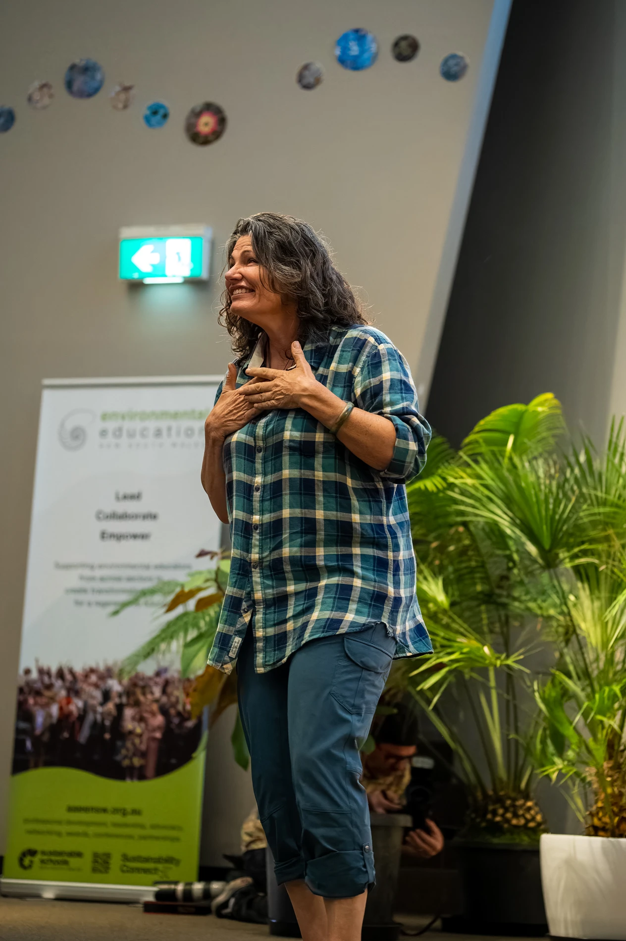 Person In Plaid Shirt, Blue Pants Gesturing Indoors, With Plants And Environmental Banner Background.