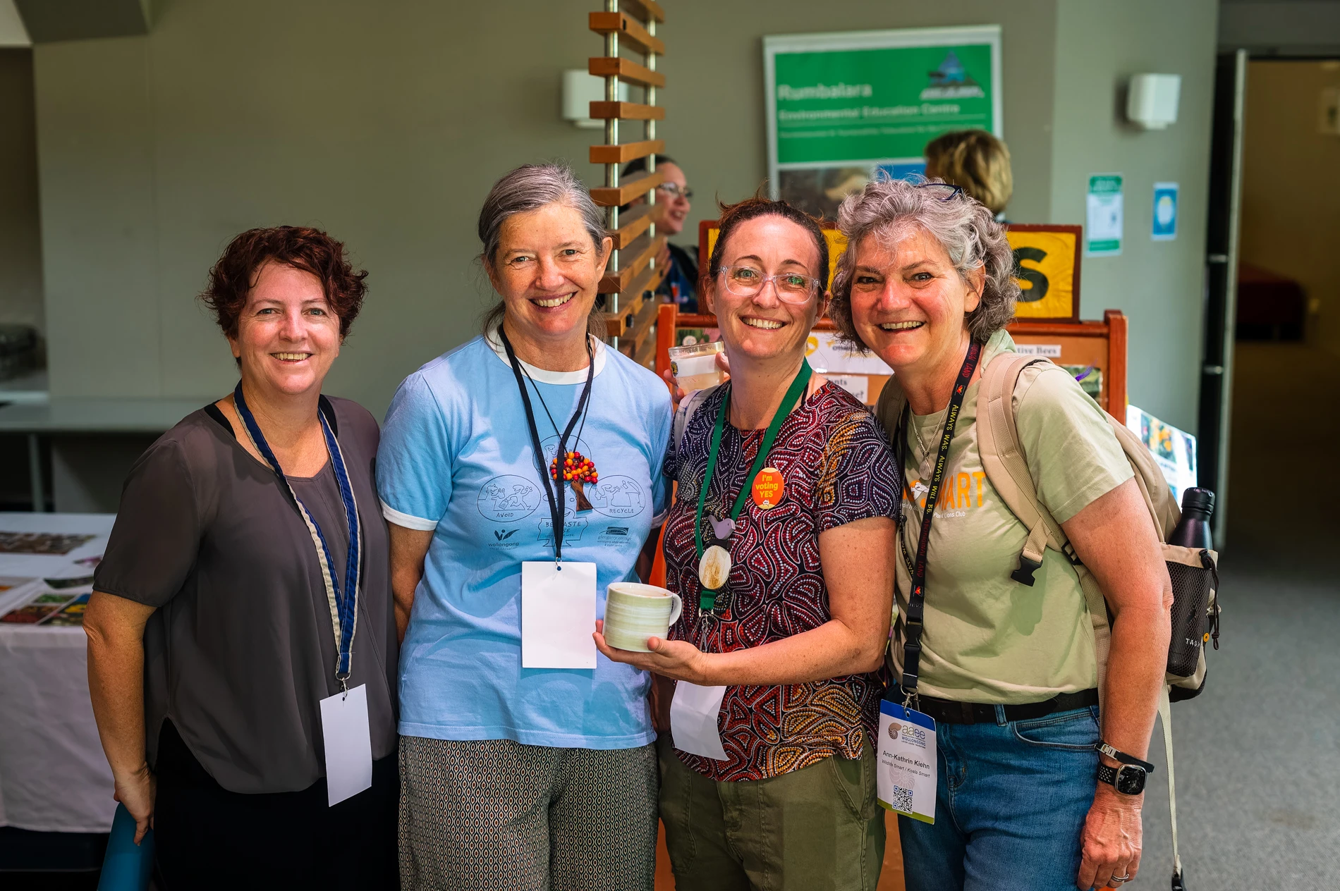 Four Women With Name Tags Smiling Indoors, One Holding A Ceramic Object, Displays In Background.