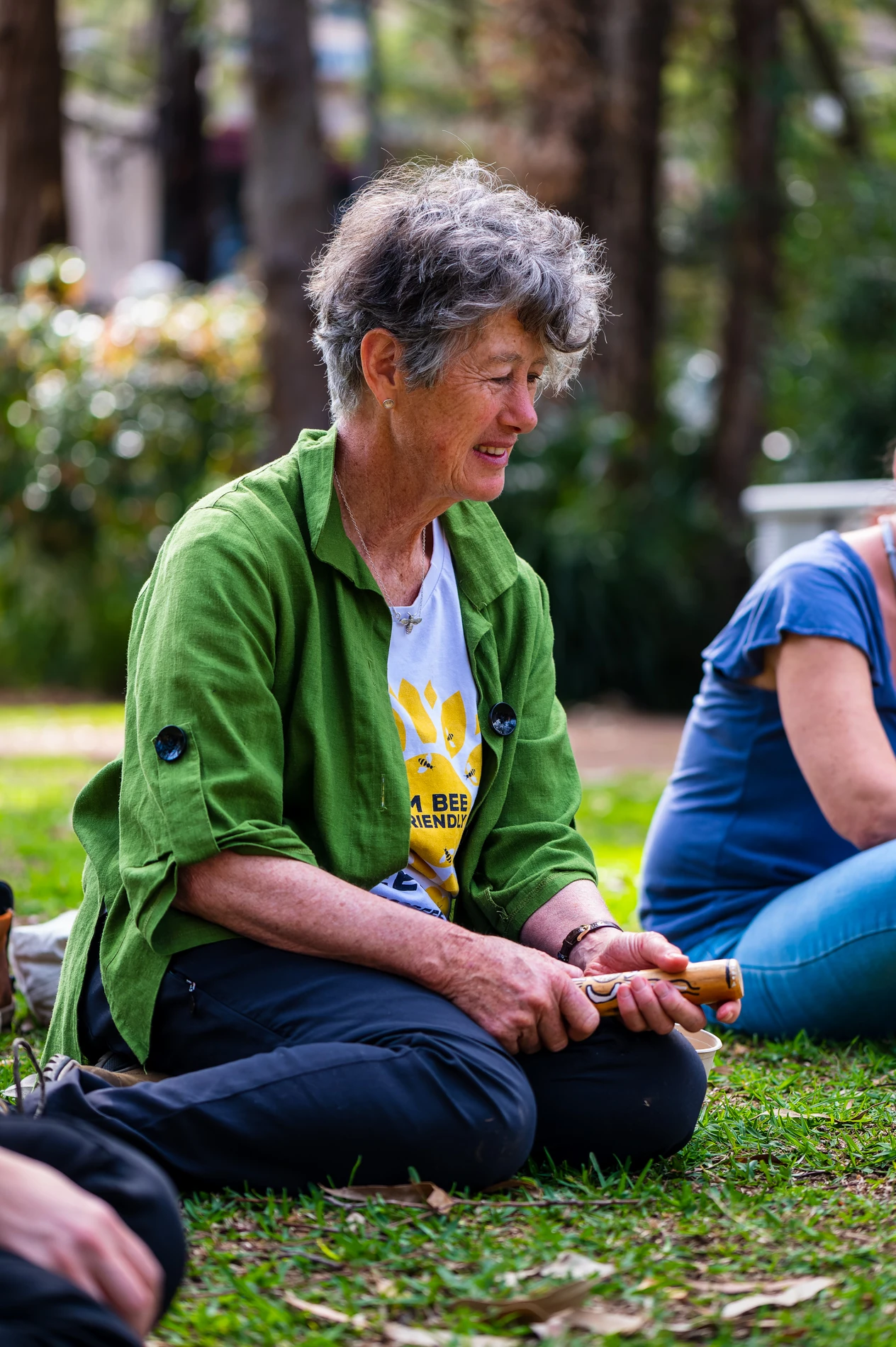 Person Sitting On Grass, Holding A Wooden Instrument, In A Green Jacket And BeeThemed TShirt.