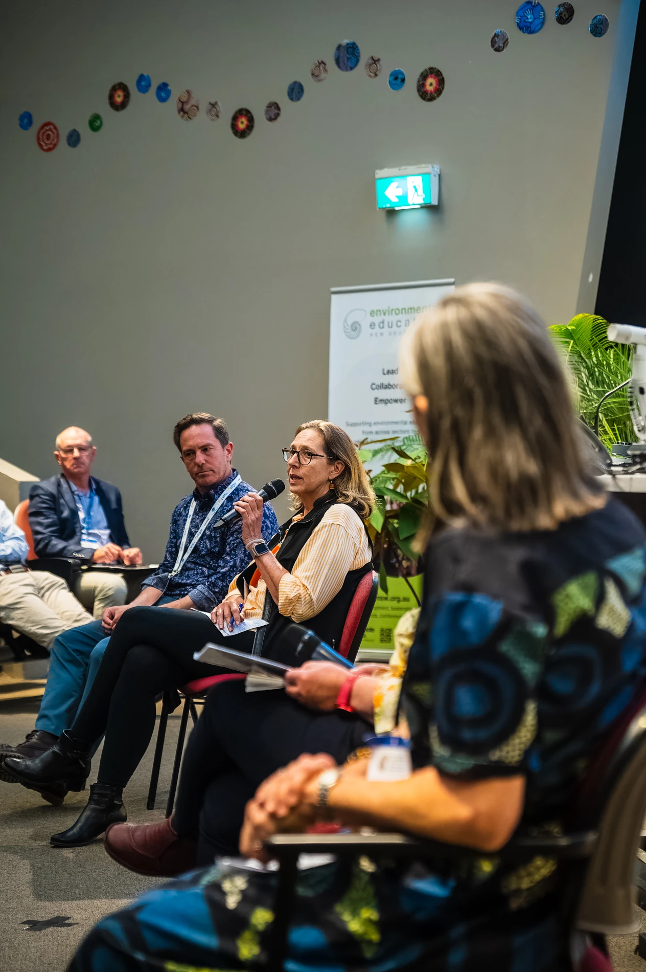 Individuals In A Panel Discussion, With A Speaker And Educational Organisation Banner In Background.