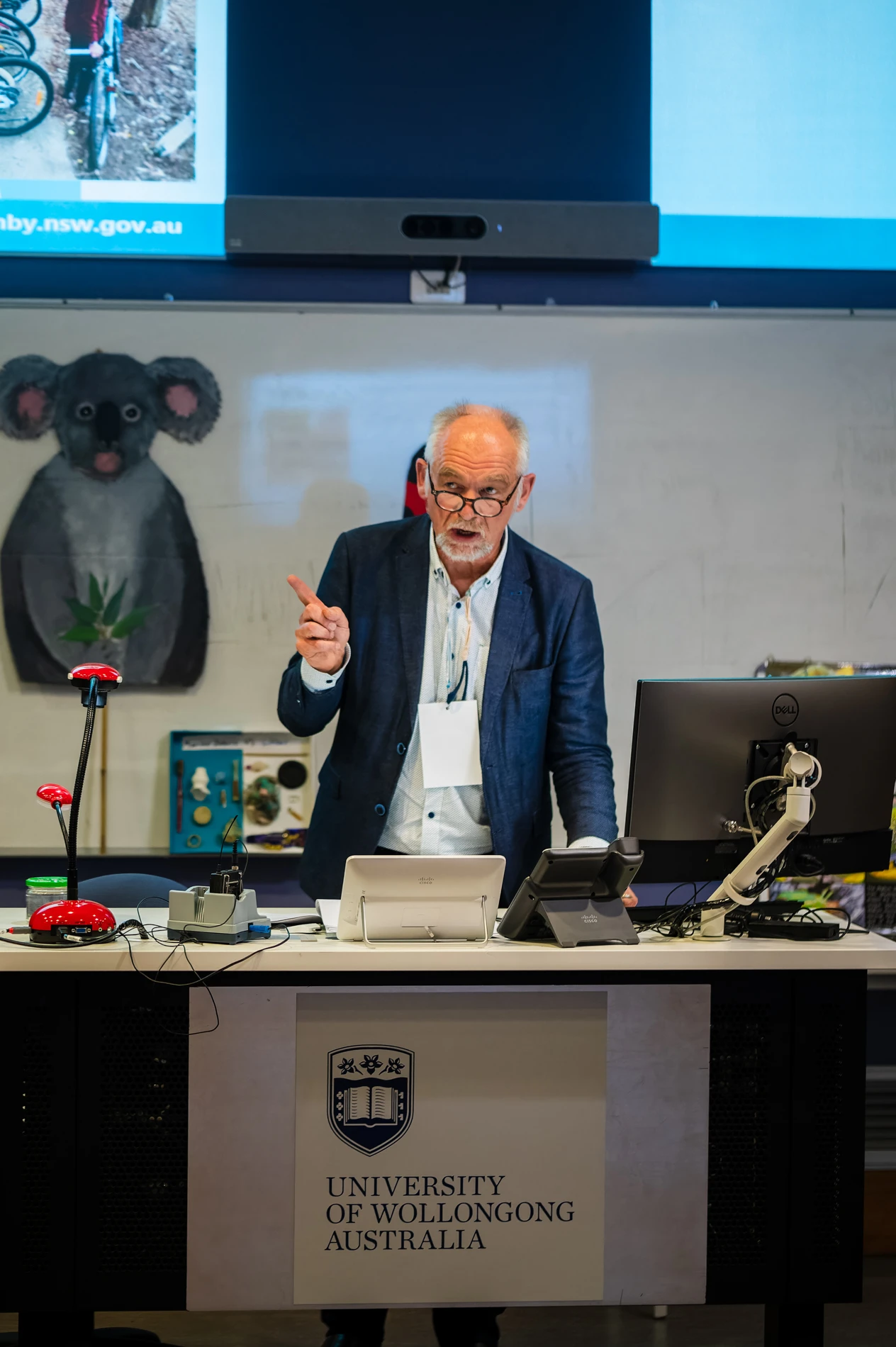 Man Speaking At University Of Wollongong Lectern With Computers And Koala Illustration.