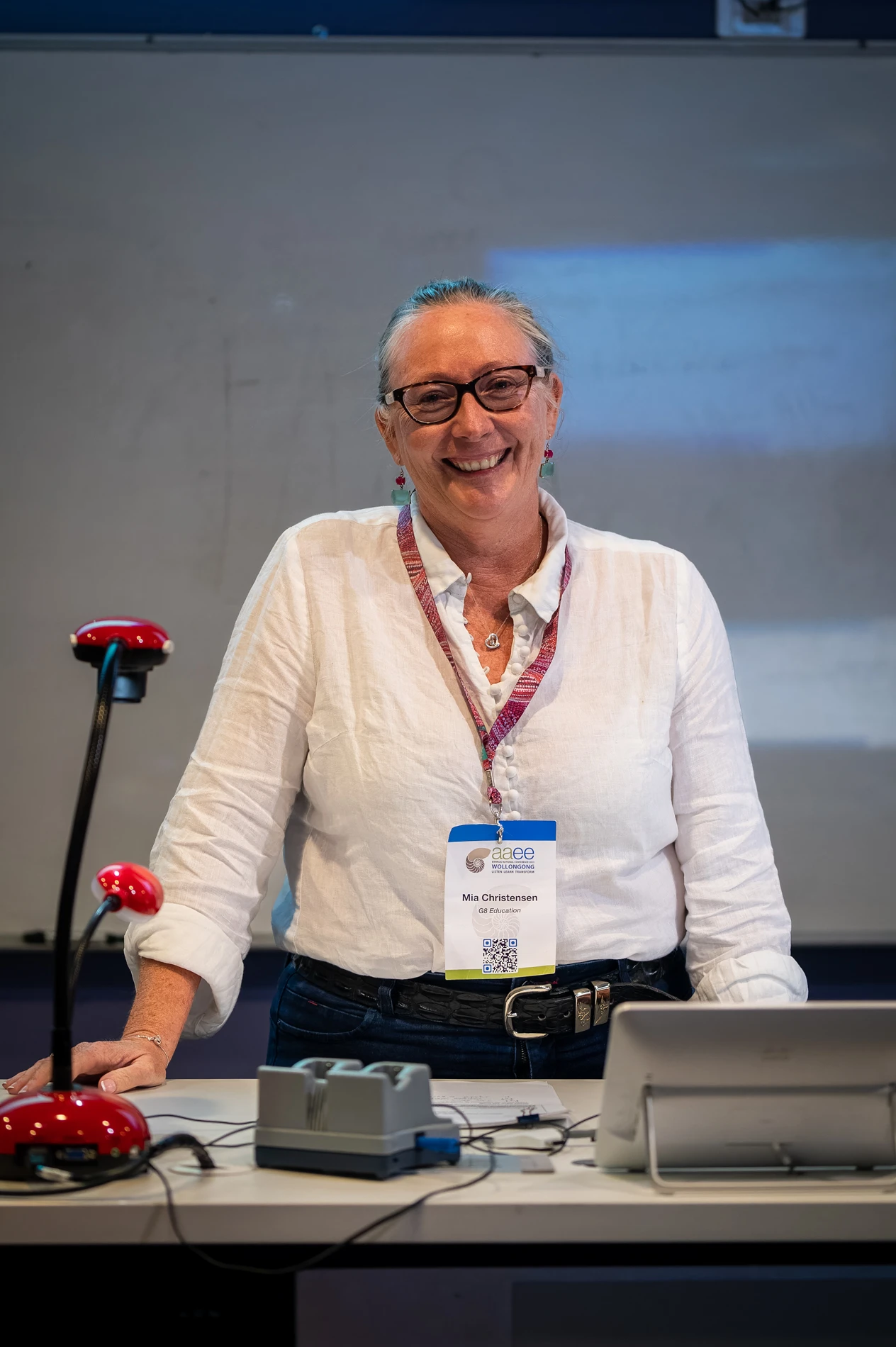Woman In Glasses With Name Badge, Smiling, At A Desk With Electronics And A Red Lamp In Front.