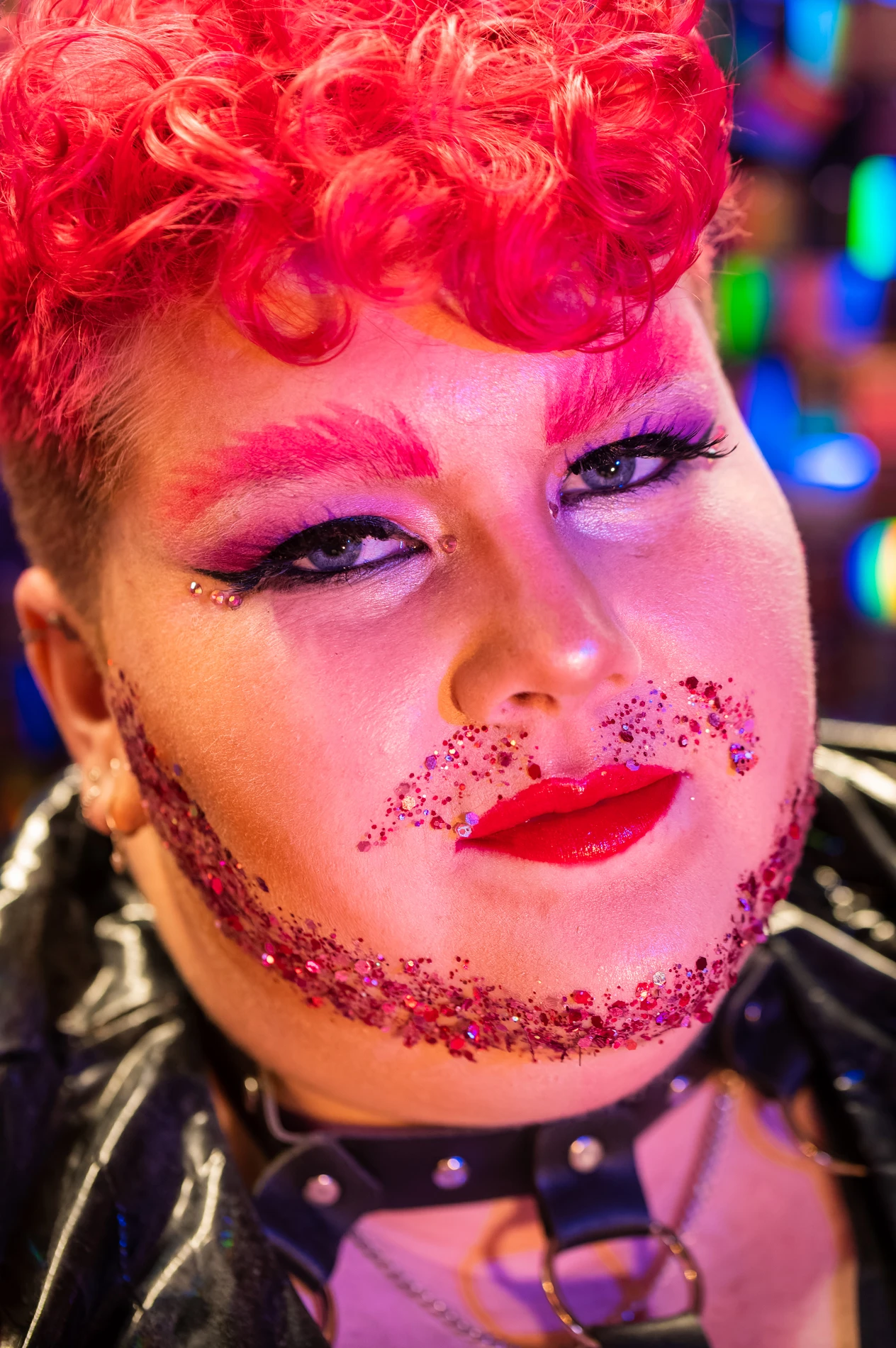 Person With Red Curly Hair, Vibrant Makeup, Glitter, Jewels, Dark Outfit, Colorful Lights Background.