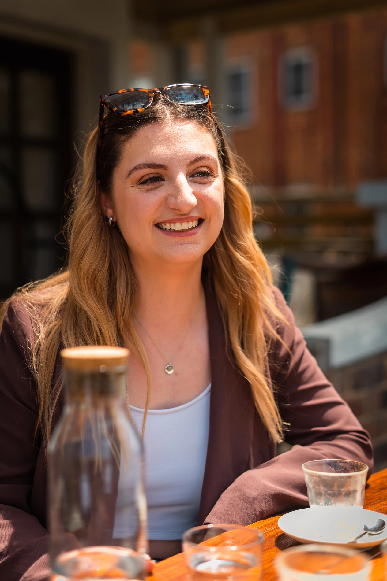 Person Smiling Outdoors With Sunglasses On Head, Brown Blazer, Glass Bottle, And Empty Dishes.