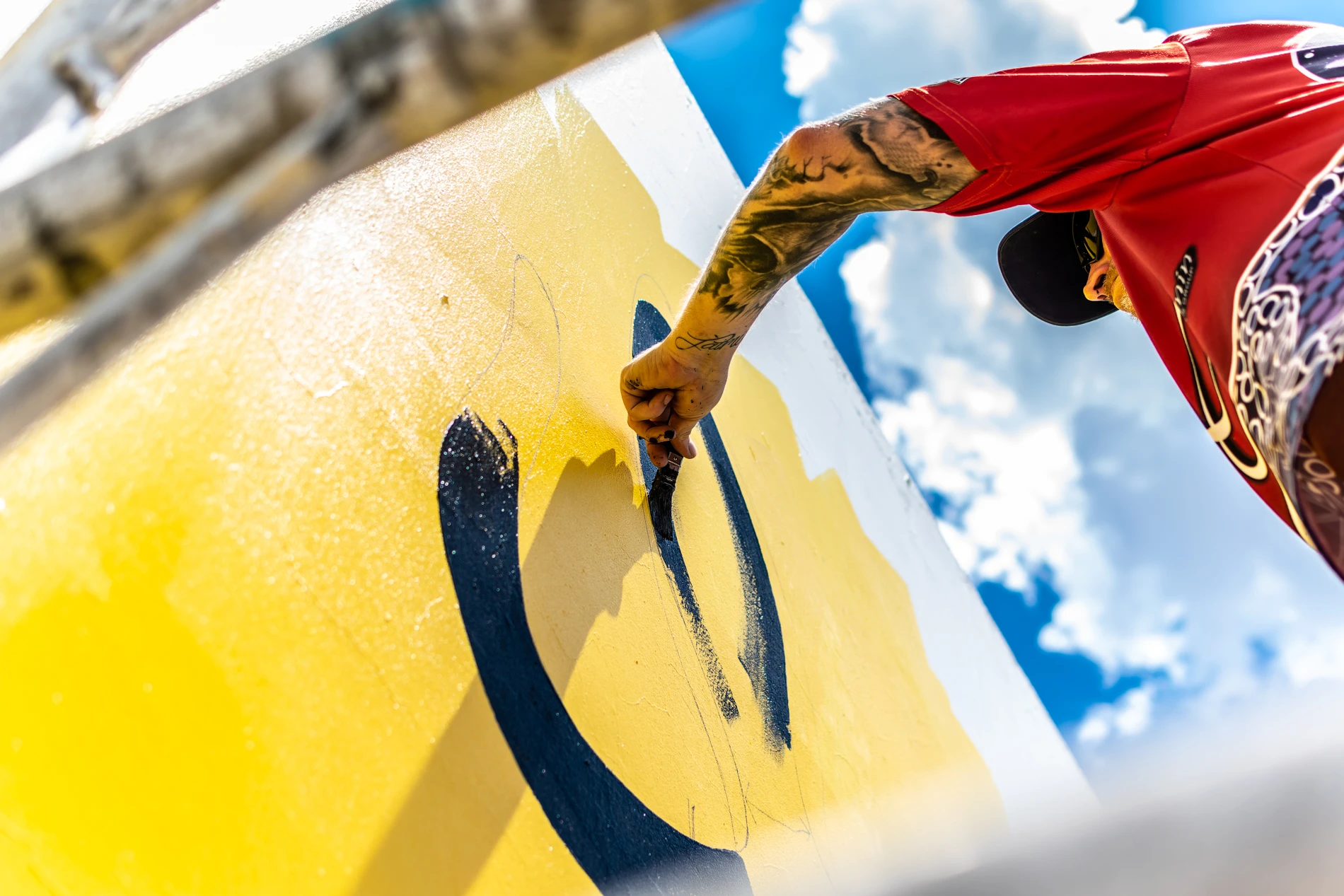 GalleriesAlleyGalleryLowAngleTattooedPainterYellowWall Person In Red Shirt Painting Yellow Mural With Dark Lines, Clear Blue Sky In Background.