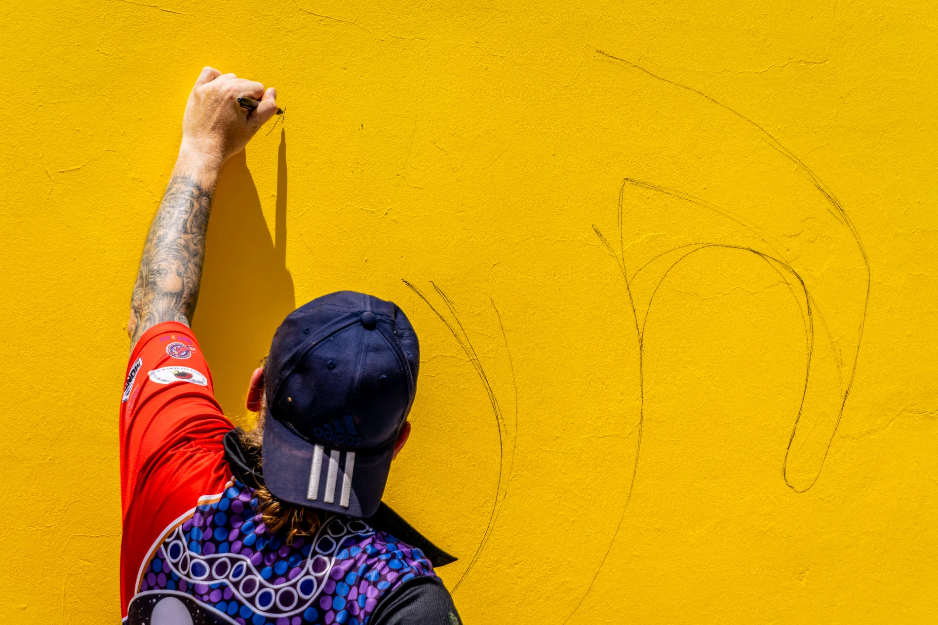 Person Sketching On A Yellow Wall Wearing A Cap And Colorful Shirt With Tattooed Arm.