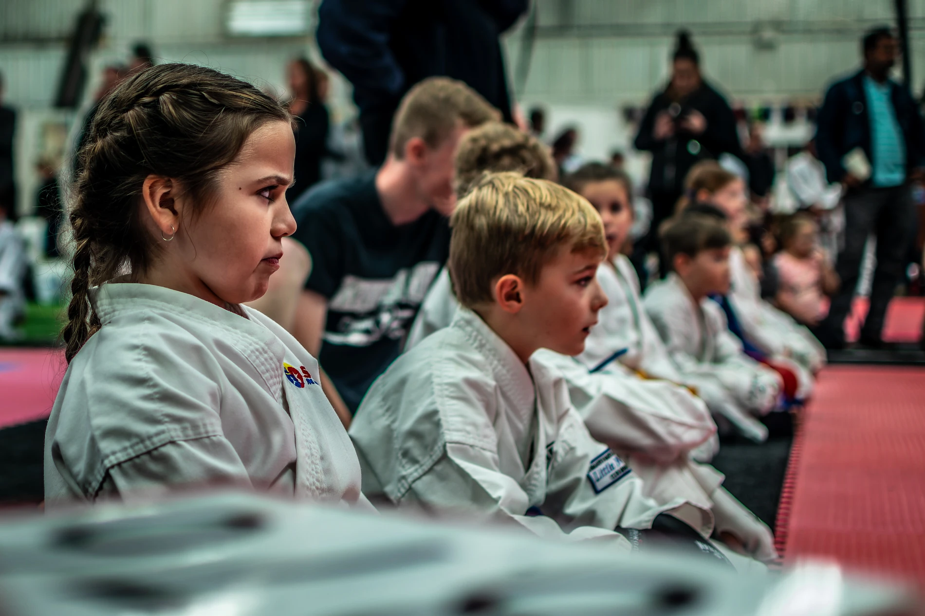 Children In Martial Arts Class, Wearing White Uniforms, With Instructors In The Background.