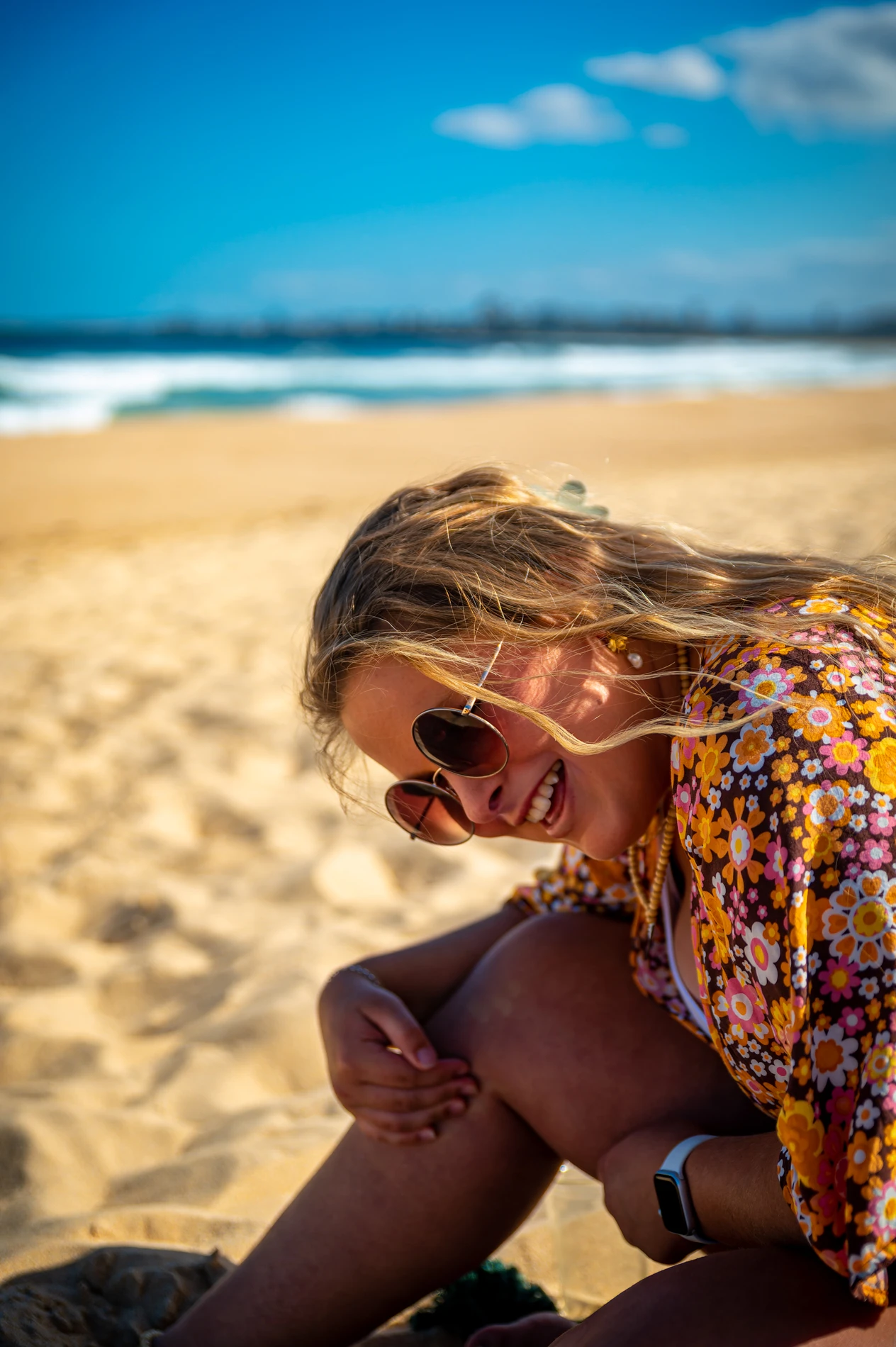 Person In Sunglasses And Floral Shirt Smiling On A Sandy Beach With Ocean In The Background.