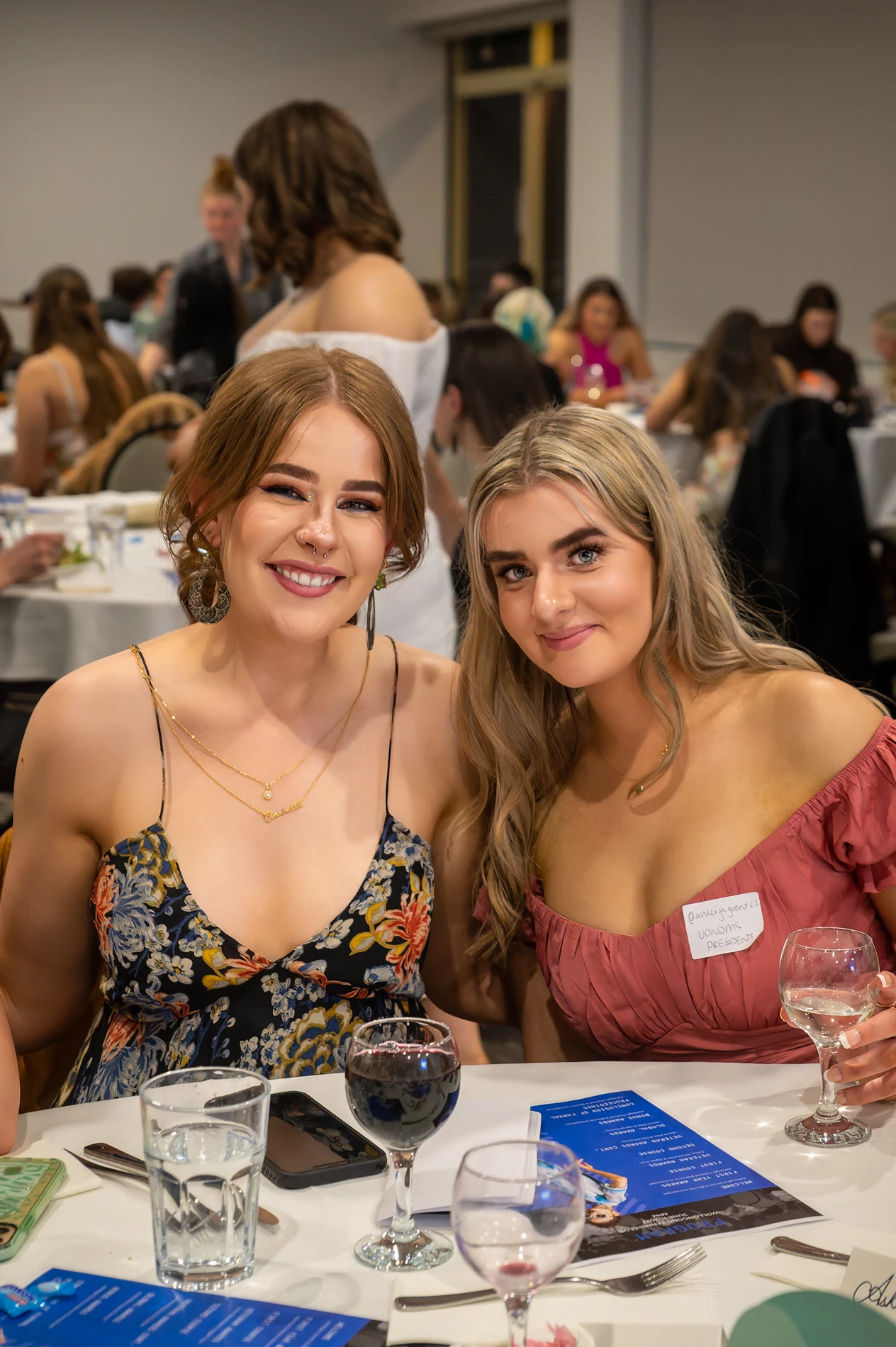 Two Women In Formal Attire Smiling At A Banquet Table With Wine, Smartphone, Menu Visible.