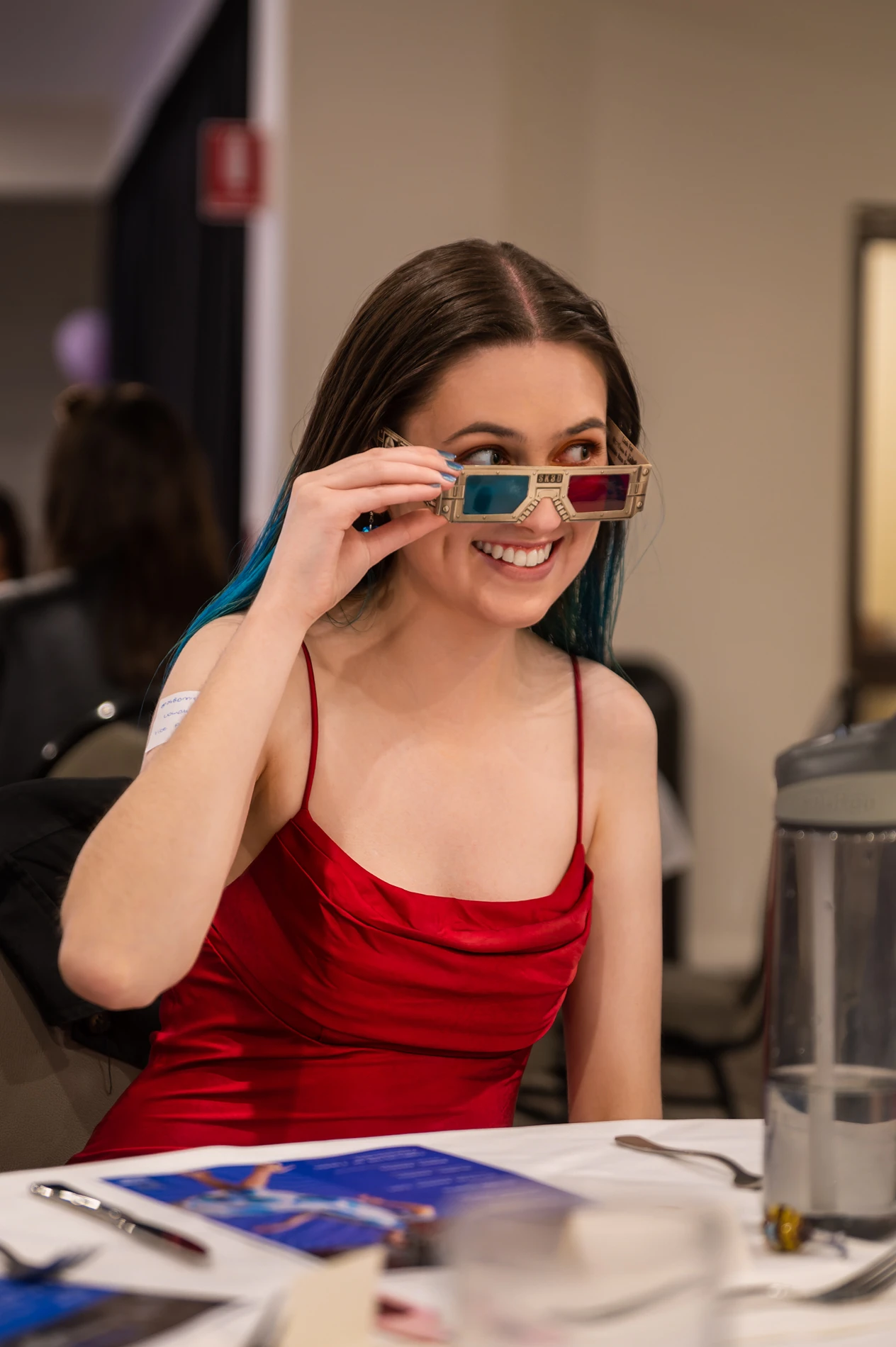 Woman In Red Dress Holds 3D Glasses, Smiling At A Table With Documents And Water Bottle.