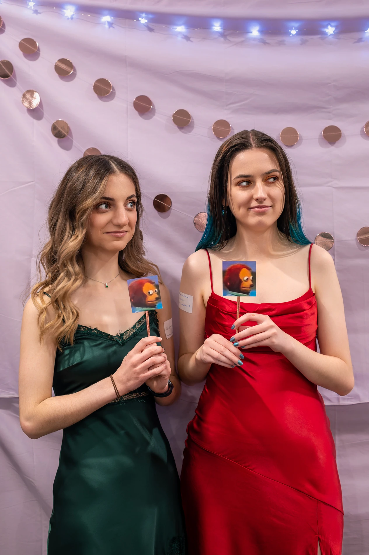 Two People In Elegant Dresses Holding Images On Sticks Against A Backdrop With Lights And Ornaments.