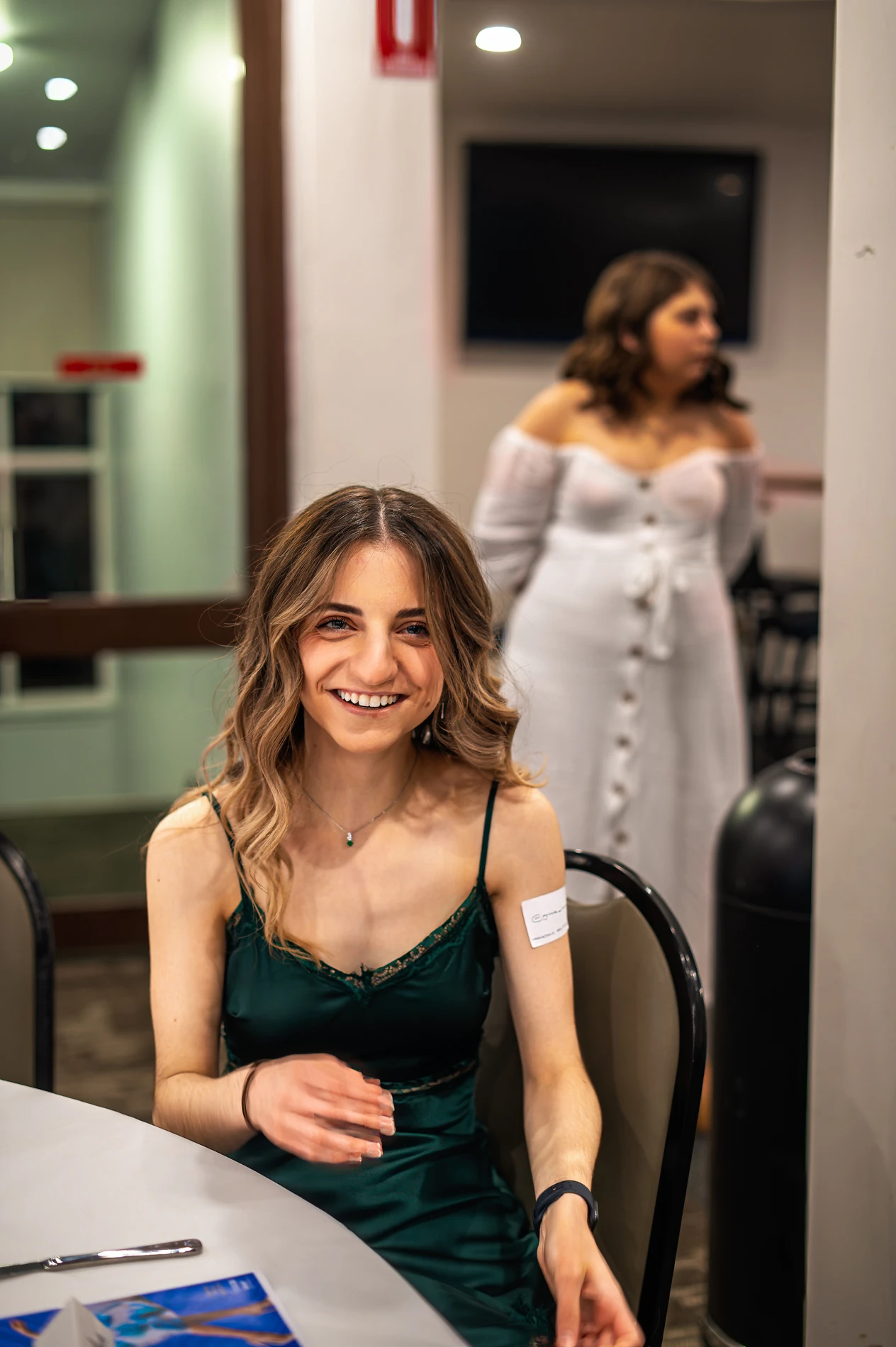 A Woman In A Green Dress Seated At A Table; Blurred Figure In White Stands Behind Indoors.