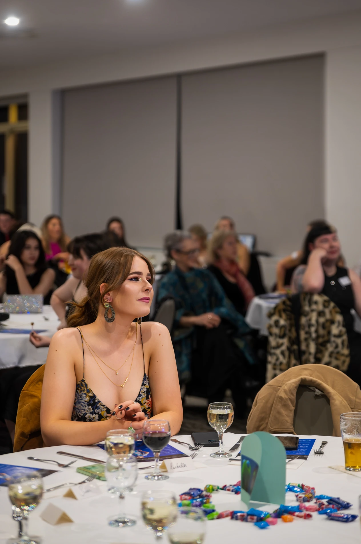 Young Woman At A Round Table Indoors, Surrounded By Event Attendees And Table Settings.