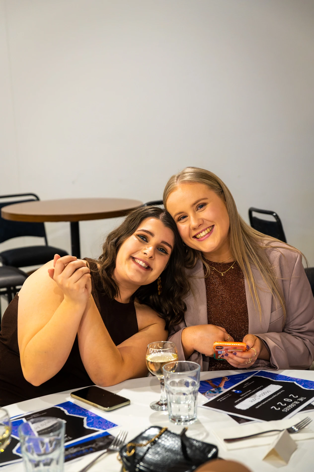 Two People Seated At A Table Indoors, Smiling With Drinks And Phone On Table.