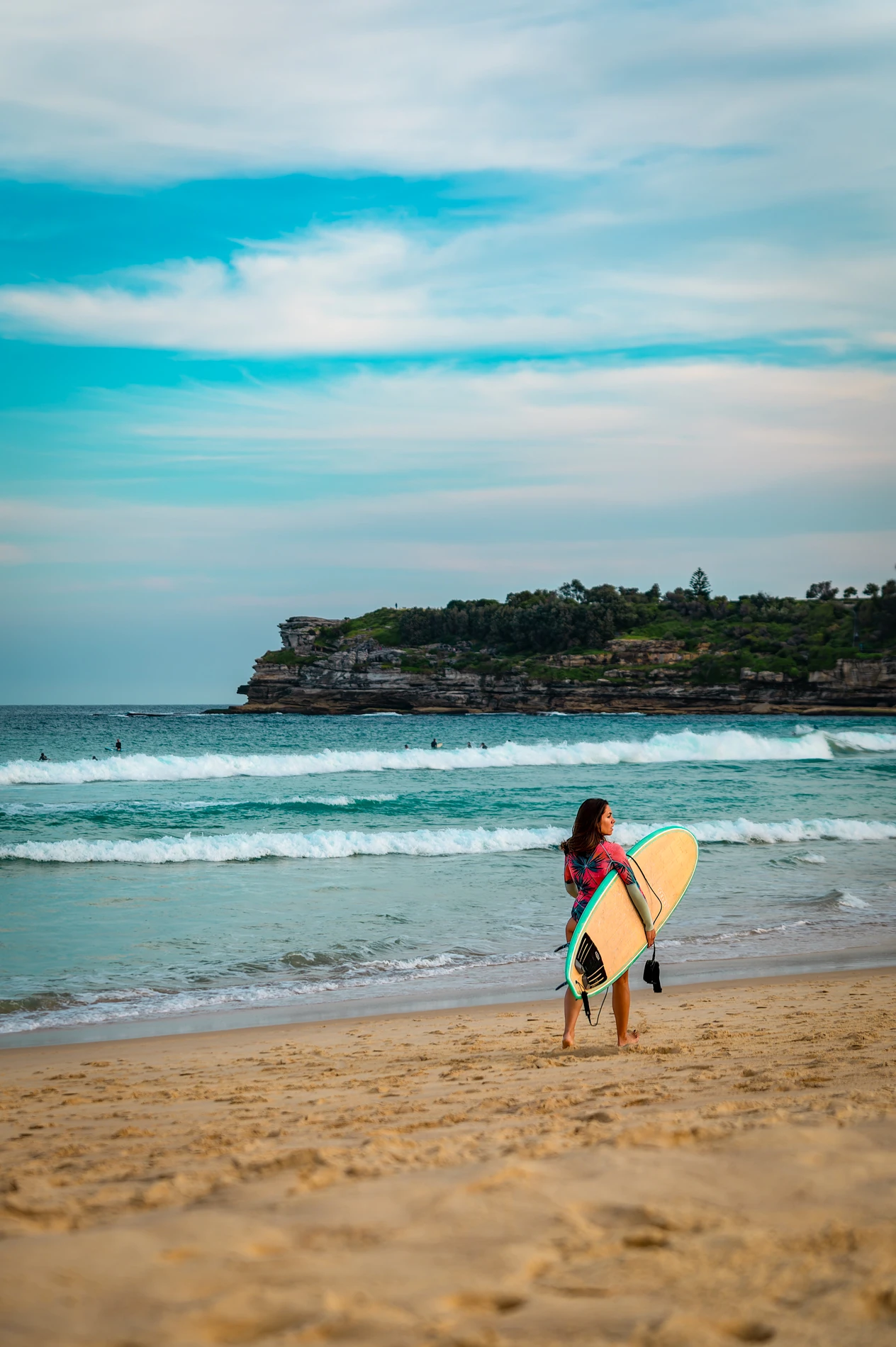 Person With Surfboard Walking On Sandy Beach Toward Ocean, With Waves And Distant Headland.