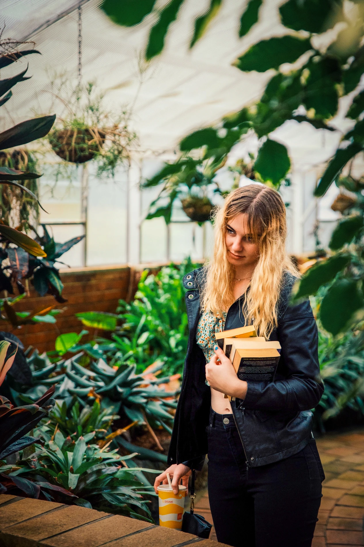 Person With Books And A Takeaway Cup In A Lush Greenhouse With Vibrant Plants.