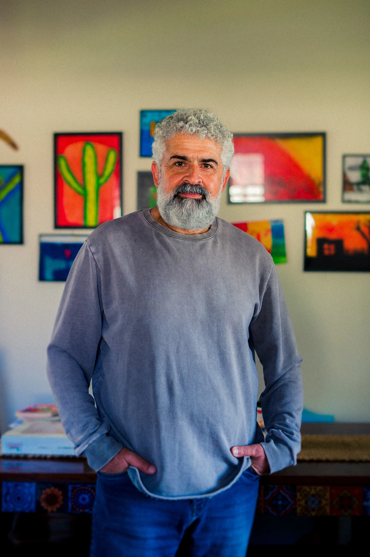 Man With Gray Hair And Beard Indoors, Wearing Casual Outfit, Colorful Artwork On Wall Behind.