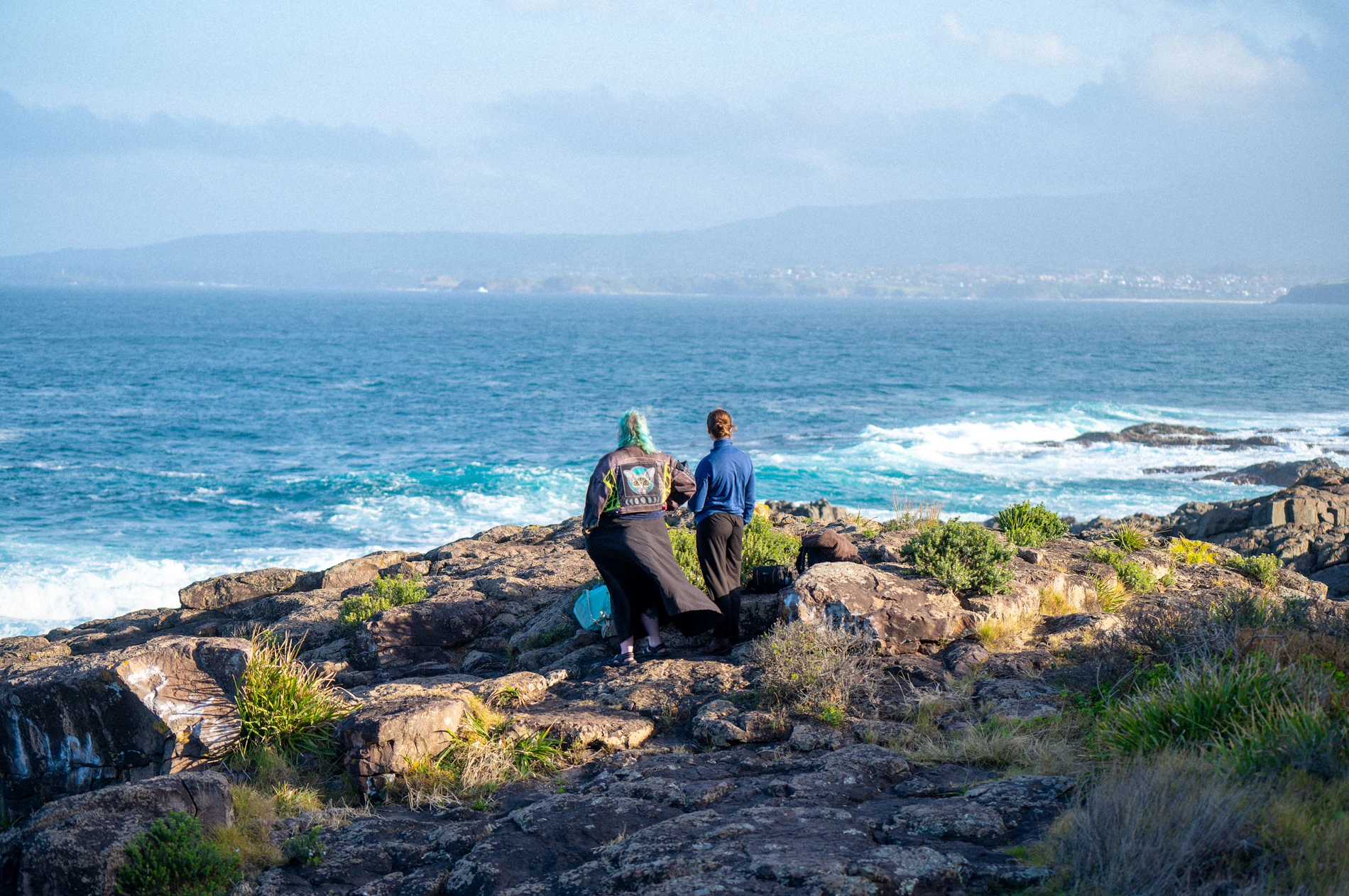 Two People On A Rocky Coast, Looking At The Ocean With Waves And Distant Hills Under A Cloudy Sky.