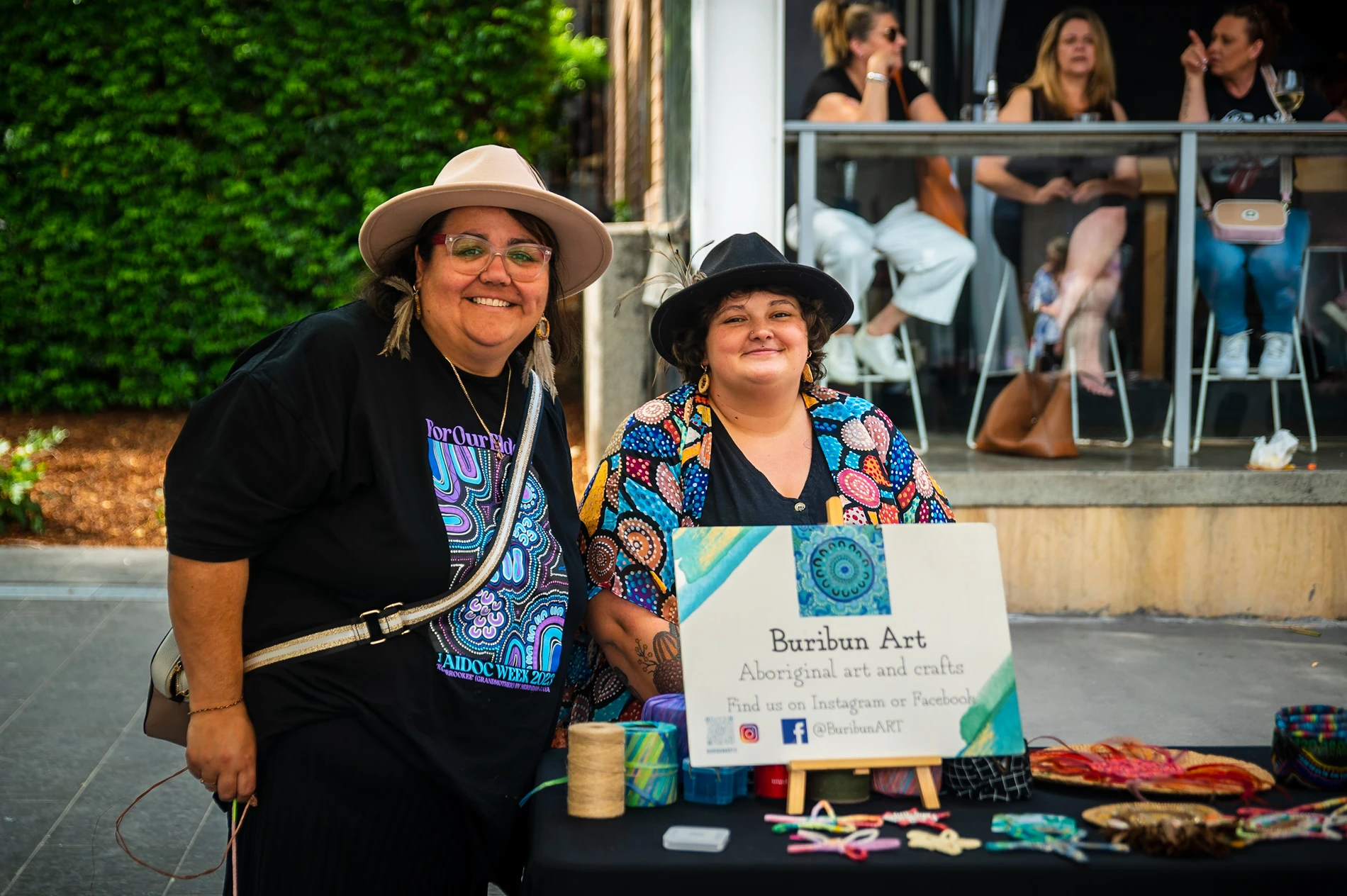 Two People At A Table With Buribun Art, Displaying Aboriginal Crafts, Outdoors Near A Cafe.