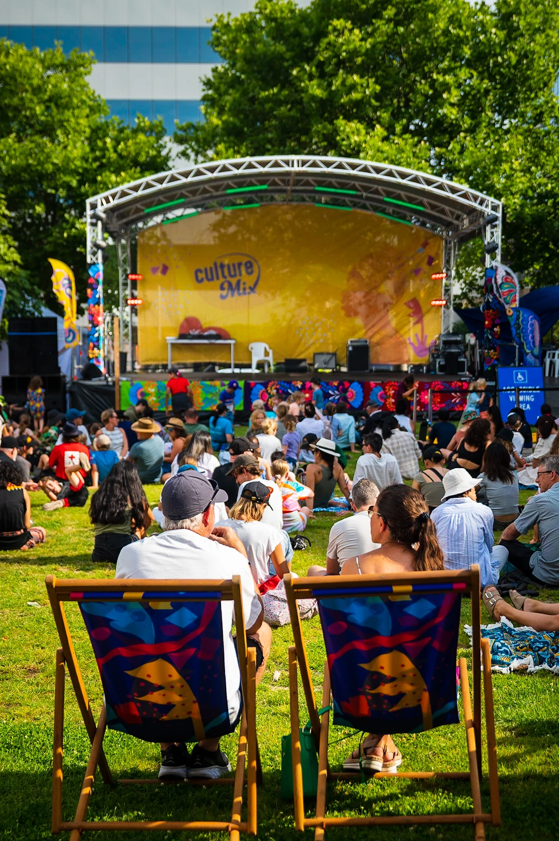 Outdoor Event With Crowd Seated On Grass, Facing A Stage With &Quot;Culture Mix&Quot; Banner, Surrounded By Trees.