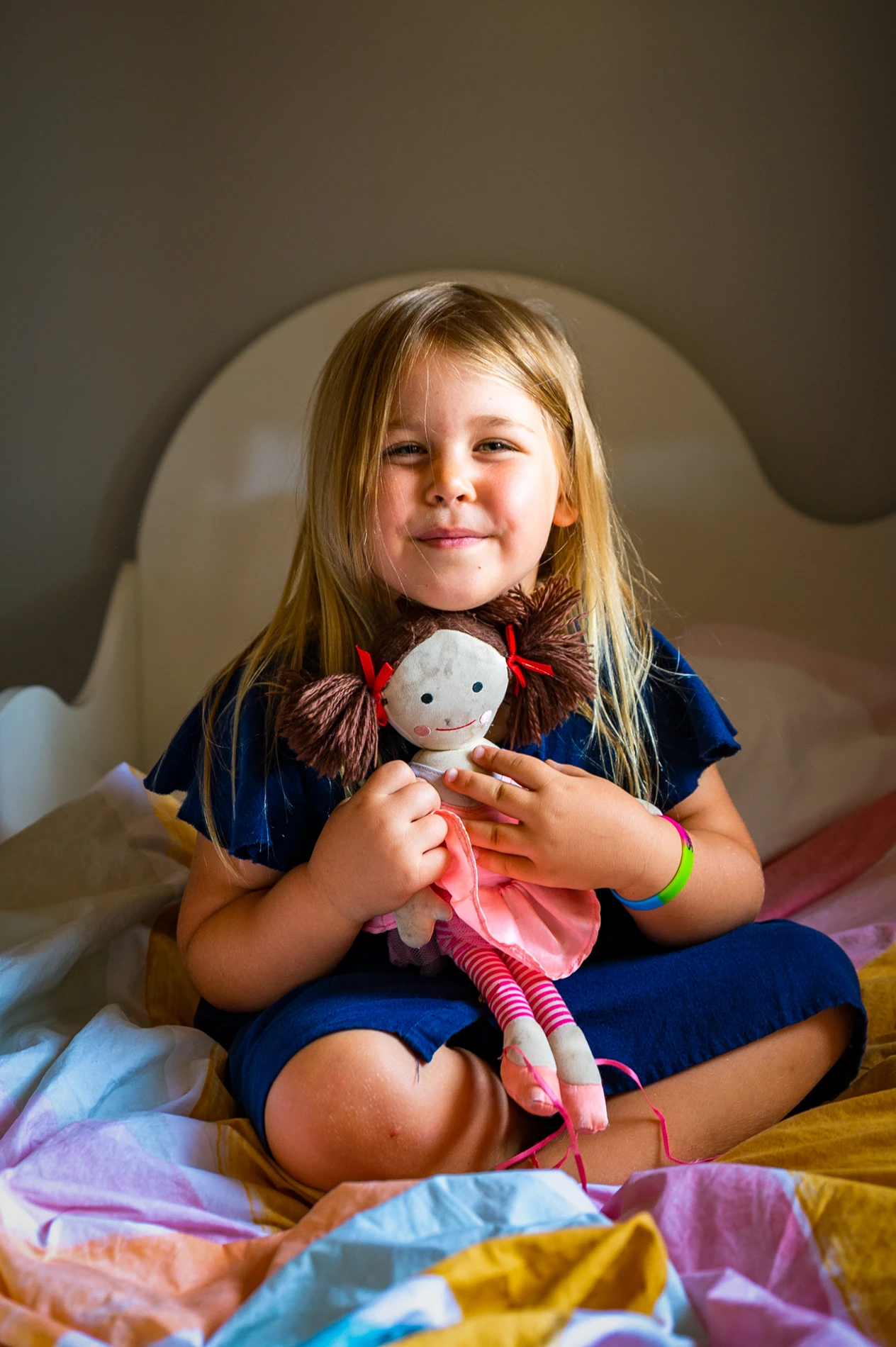 Young Girl With Blond Hair Holding A Doll On A Colorful Bed In Softly Lit Indoor Setting.