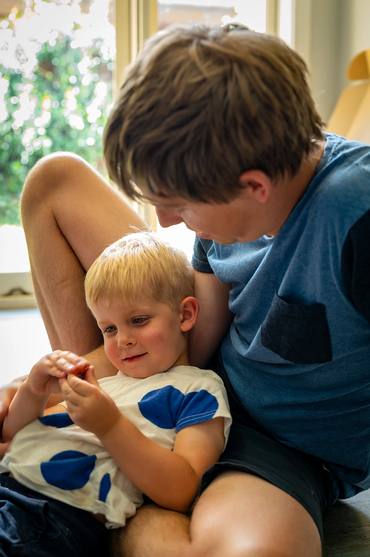 Man And Child Sitting Indoors, Child Holding Small Object, Both Appear Relaxed.
