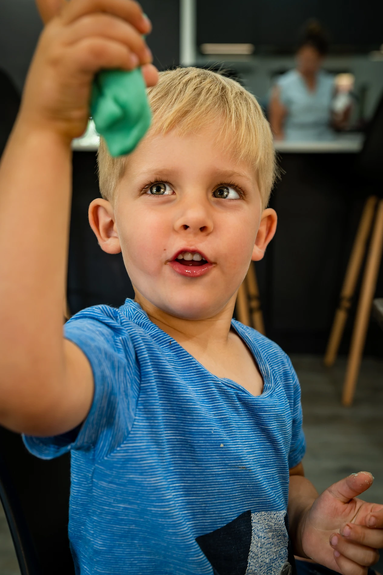 Young Child In Blue Shirt Holding A Green Object Indoors, Person Blurred In Kitchen Background.