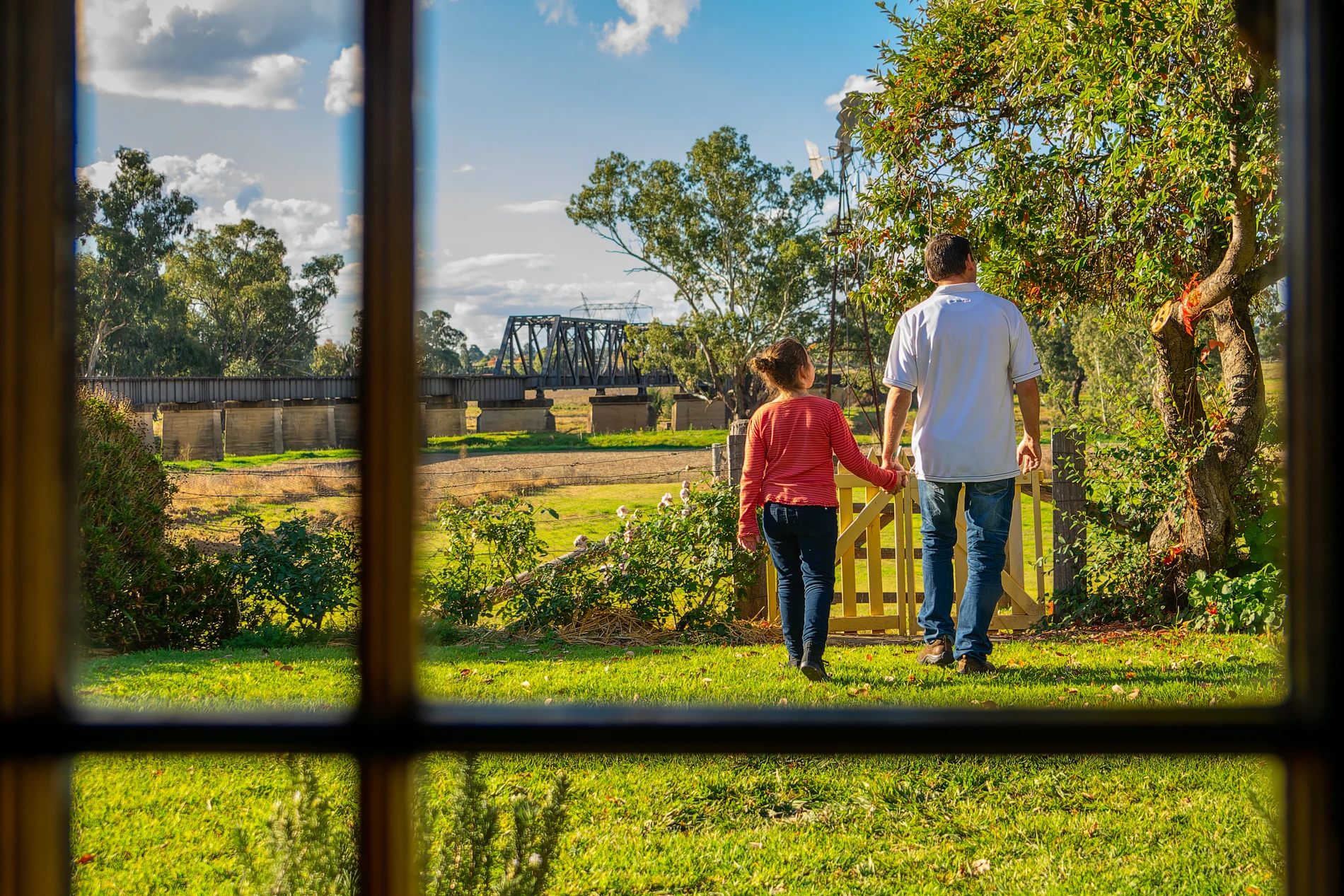 Person And Child Walking Hand In Hand Through Garden Gate Towards Landscape Seen Through Window.