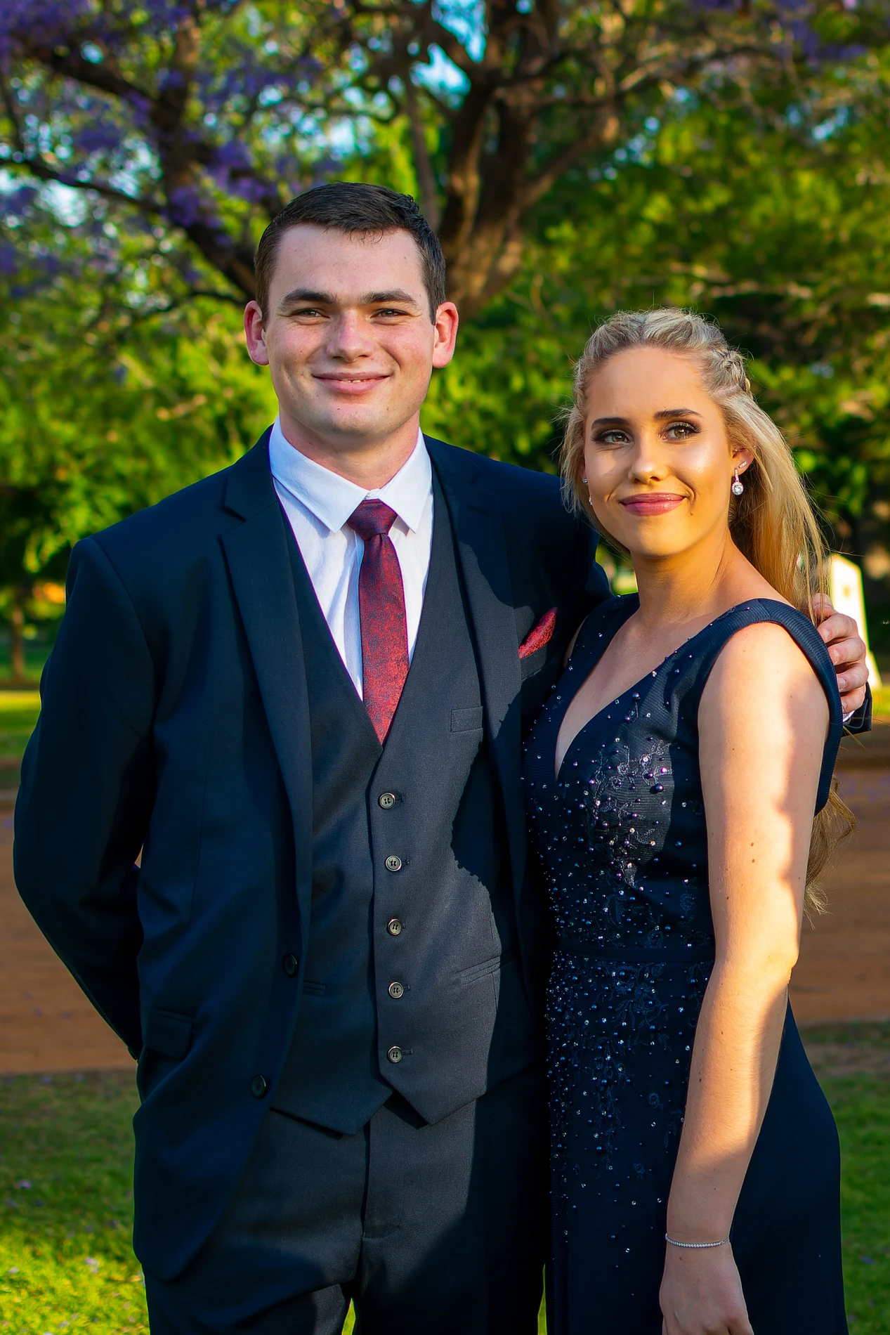 Formal Couple Standing Outdoors, Man In Dark Suit And Maroon Tie, Woman In Embellished Dress.