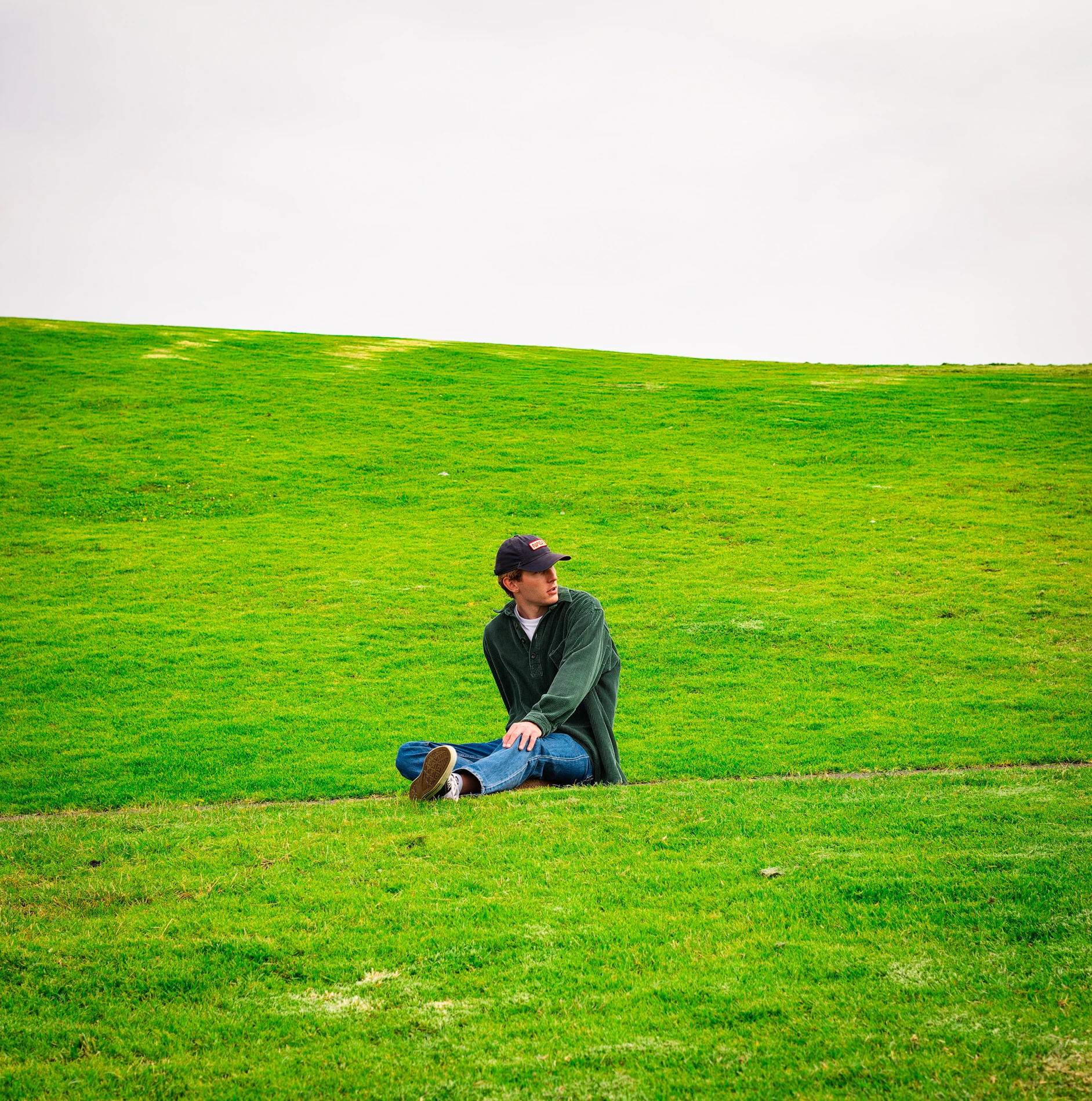 Person In Cap, Dark Jacket, And Jeans Sitting Cross-Legged On A Green Hill Under An Overcast Sky.