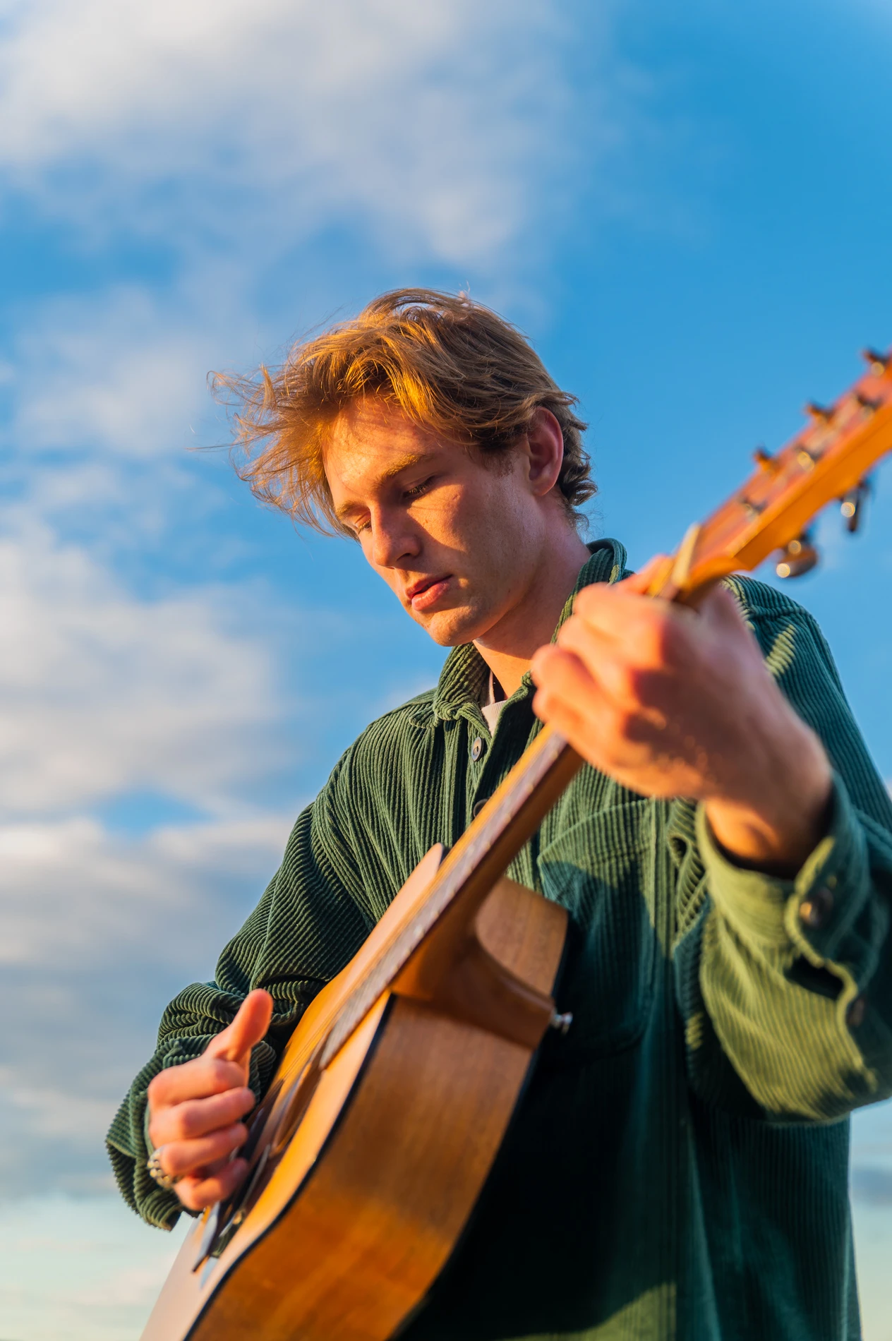 Person With Light Brown Hair Plays Acoustic Guitar Outdoors Under A Clear Blue Sky.