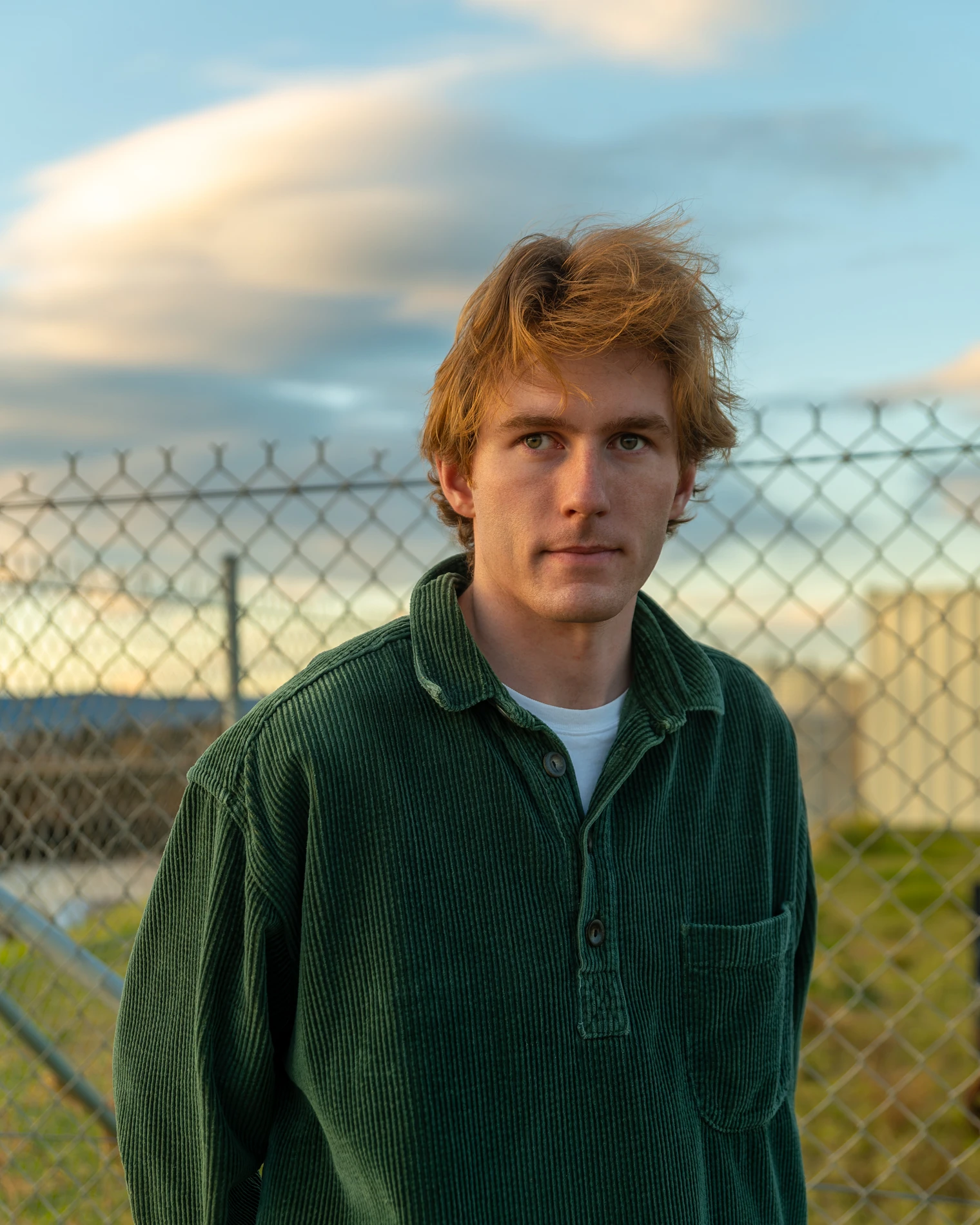 Young Man In Green Corduroy Shirt By Chain-Link Fence With Cloudy Sky Background.