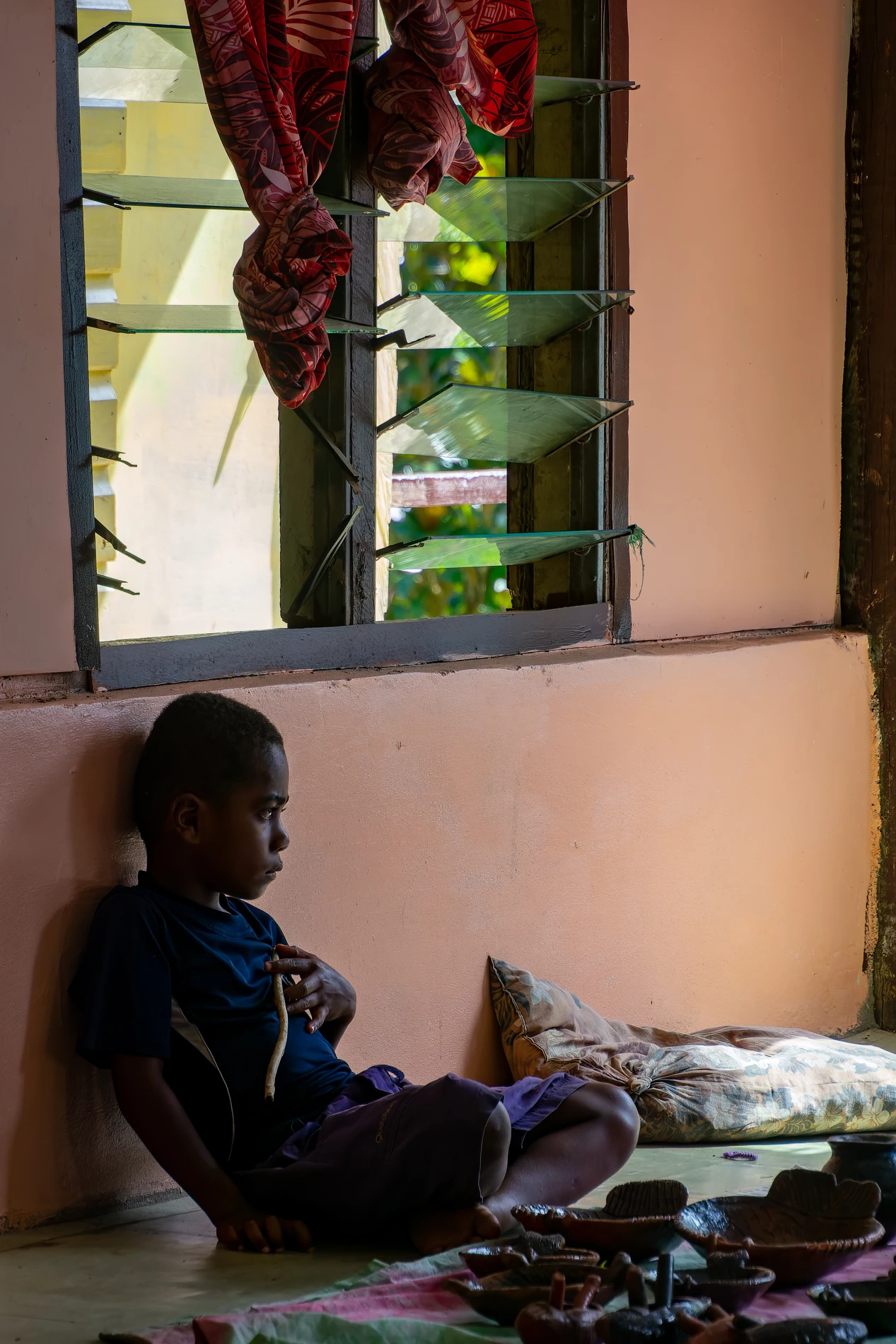 Young Boy Indoors, Leaning On Wall With Stick And Clay Objects Around, Near Window With Shutters.