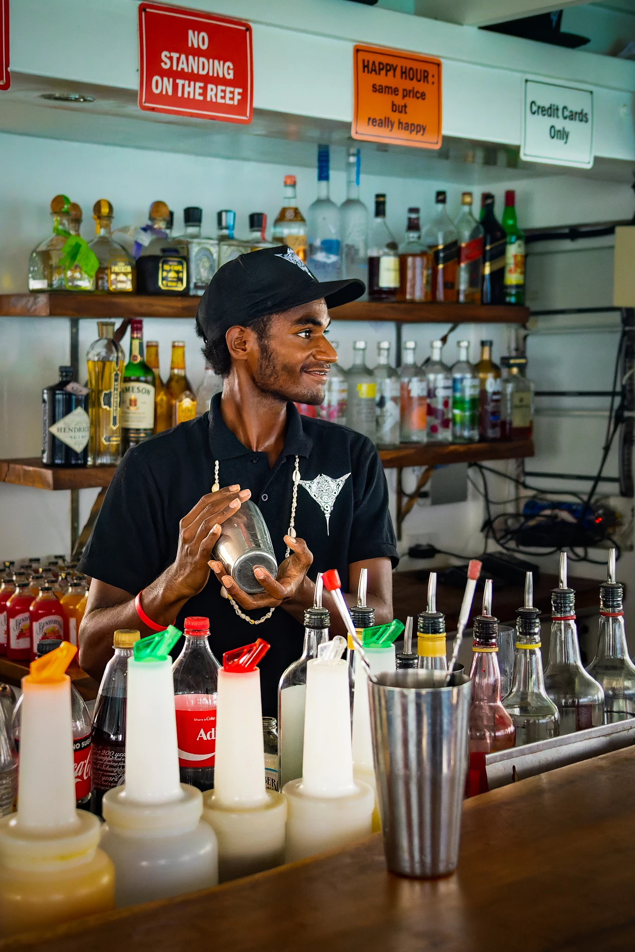 Bartender In Black Shirt Shaking Cocktail Mixer At A Well-Stocked Bar With Signs Above.