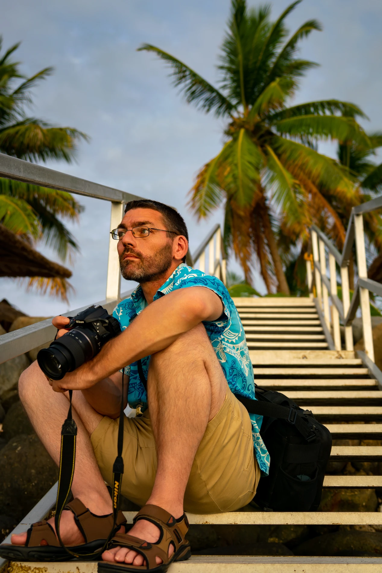 Man In Blue Shirt, Khaki Shorts, Holding Dslr On Outdoor Stairs With Palm Trees In Background.