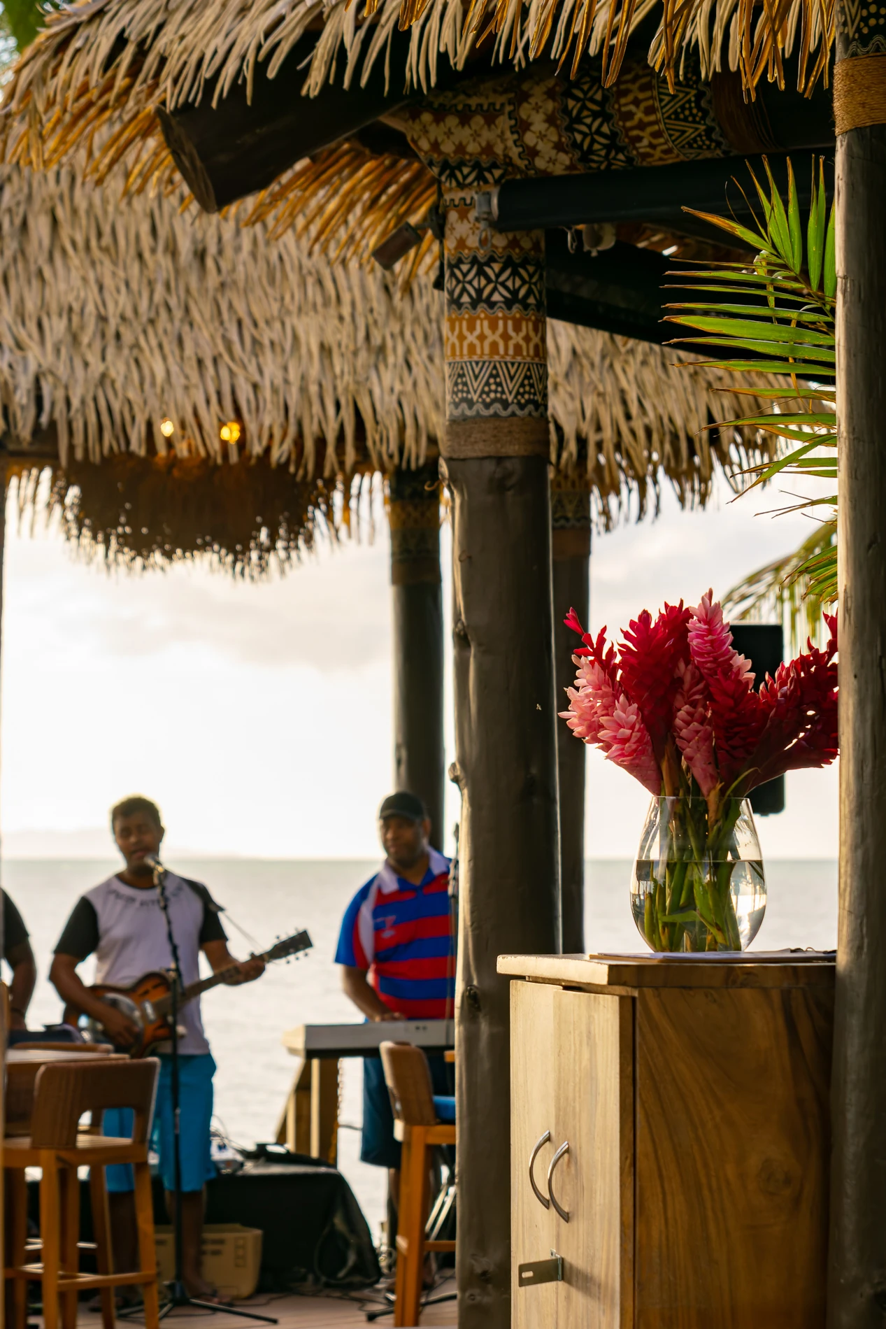 Tropical Venue With Thatched Roof, Red Flowers, And Musicians Performing By The Sea.