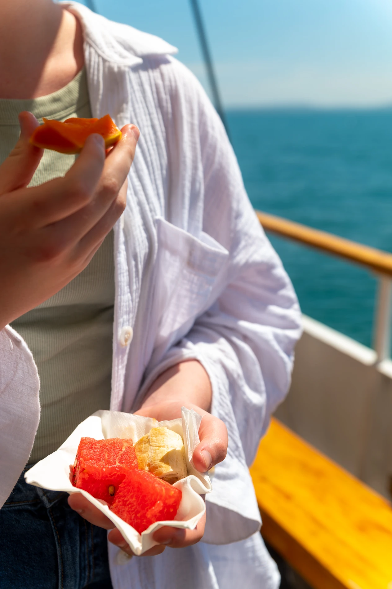 Person In White Shirt On Boat Holding Watermelon And Bananas, With One Hand Raised To Mouth.