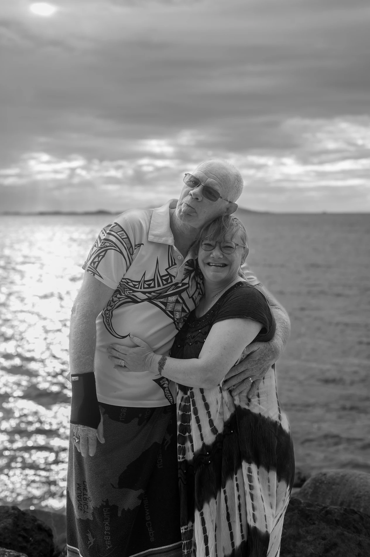 Elderly Couple Embraces By The Seaside, Ocean And Cloudy Sky In Background, Black And White.