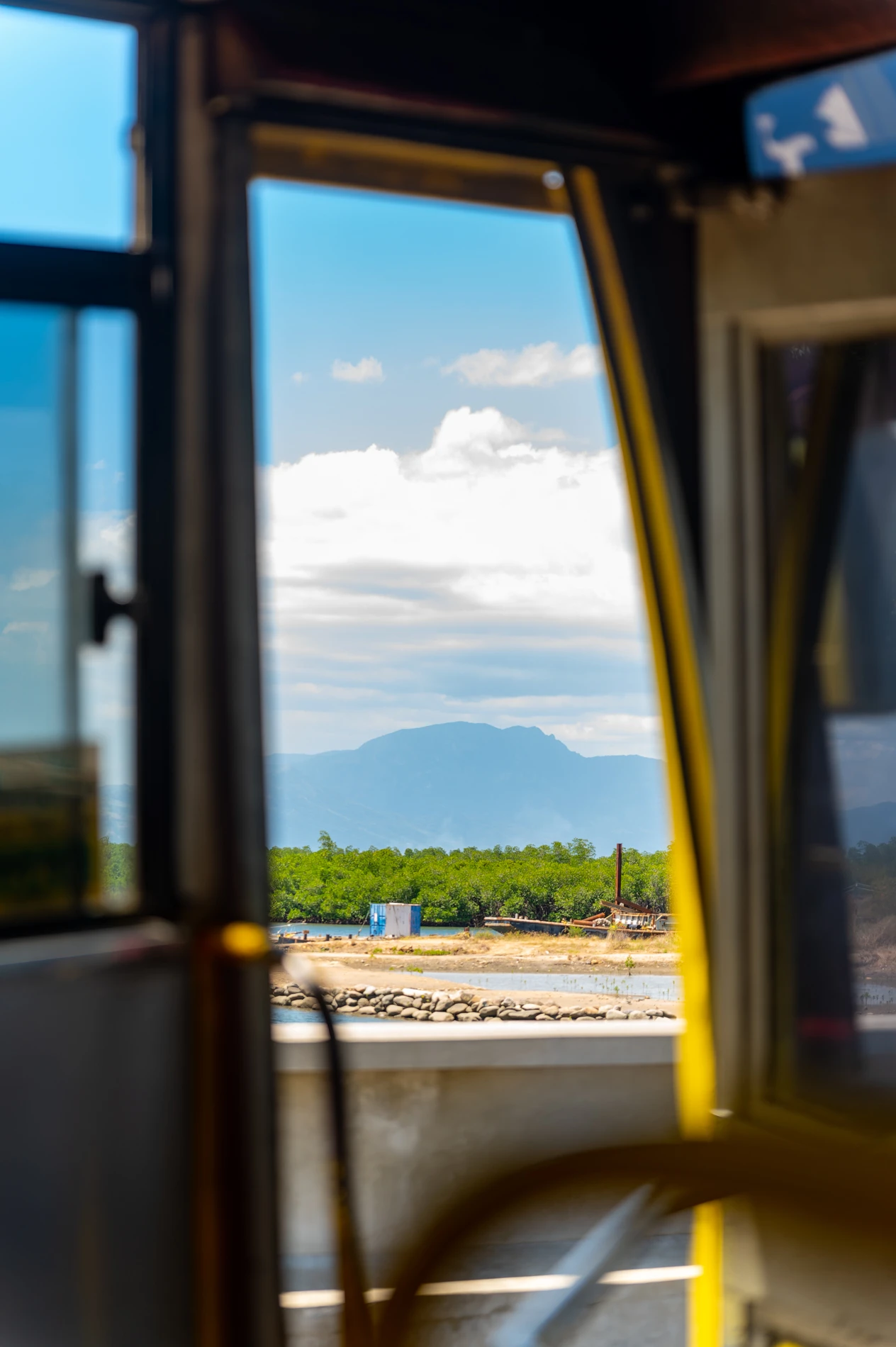 View Through An Open Window Of A Shoreline, Greenery, And Distant Mountain Under A Cloudy Sky.