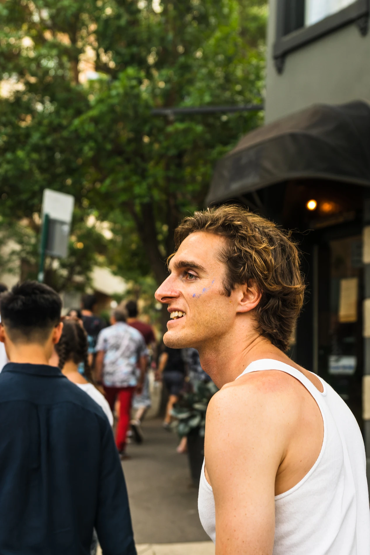 Man In White Tank Top Smiling In Outdoor Pedestrian Area With Greenery In Background.