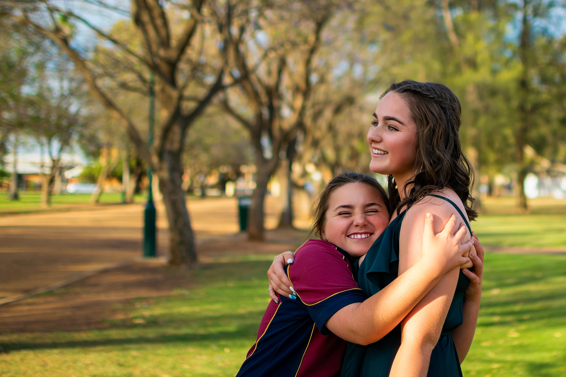 Two People Embracing In A Sunny Park, One In A Green Dress, The Other In A Red And Blue Shirt.