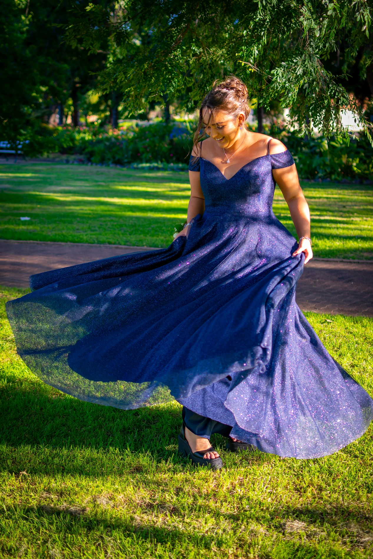 Woman Twirling In A Sparkling Navy Gown In A Sunny Park With Trees And A Path Behind Her.